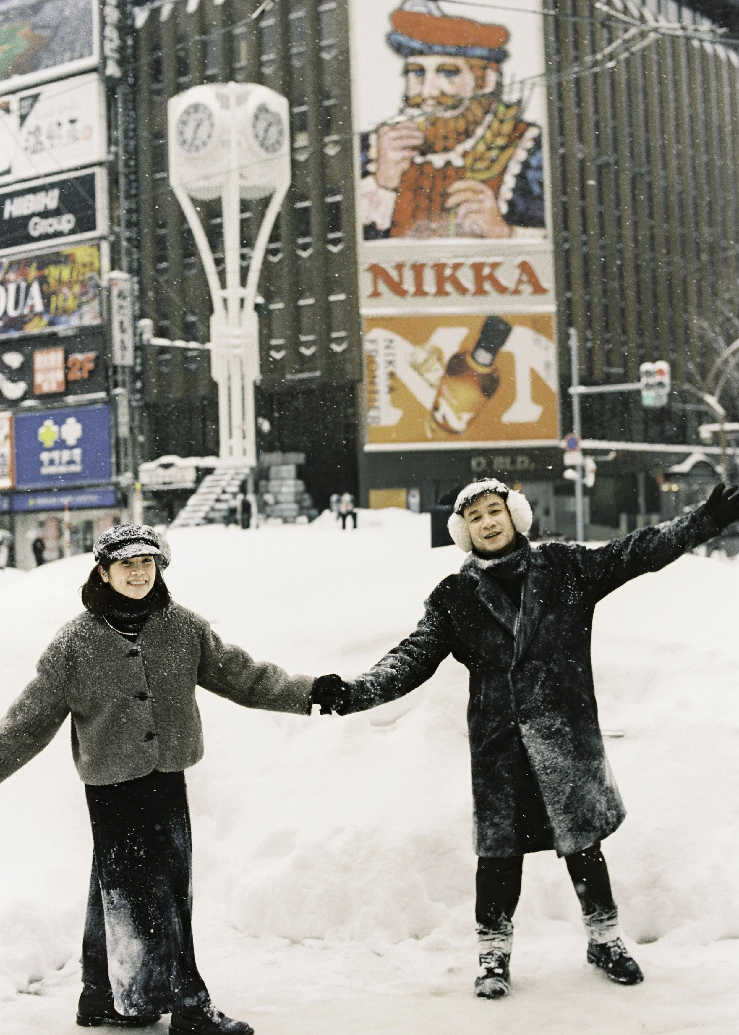 Two people holding hands in the snow in an urban area with billboards and signs in the background, wearing winter clothing. Couple photoshoot in Sapporo.
