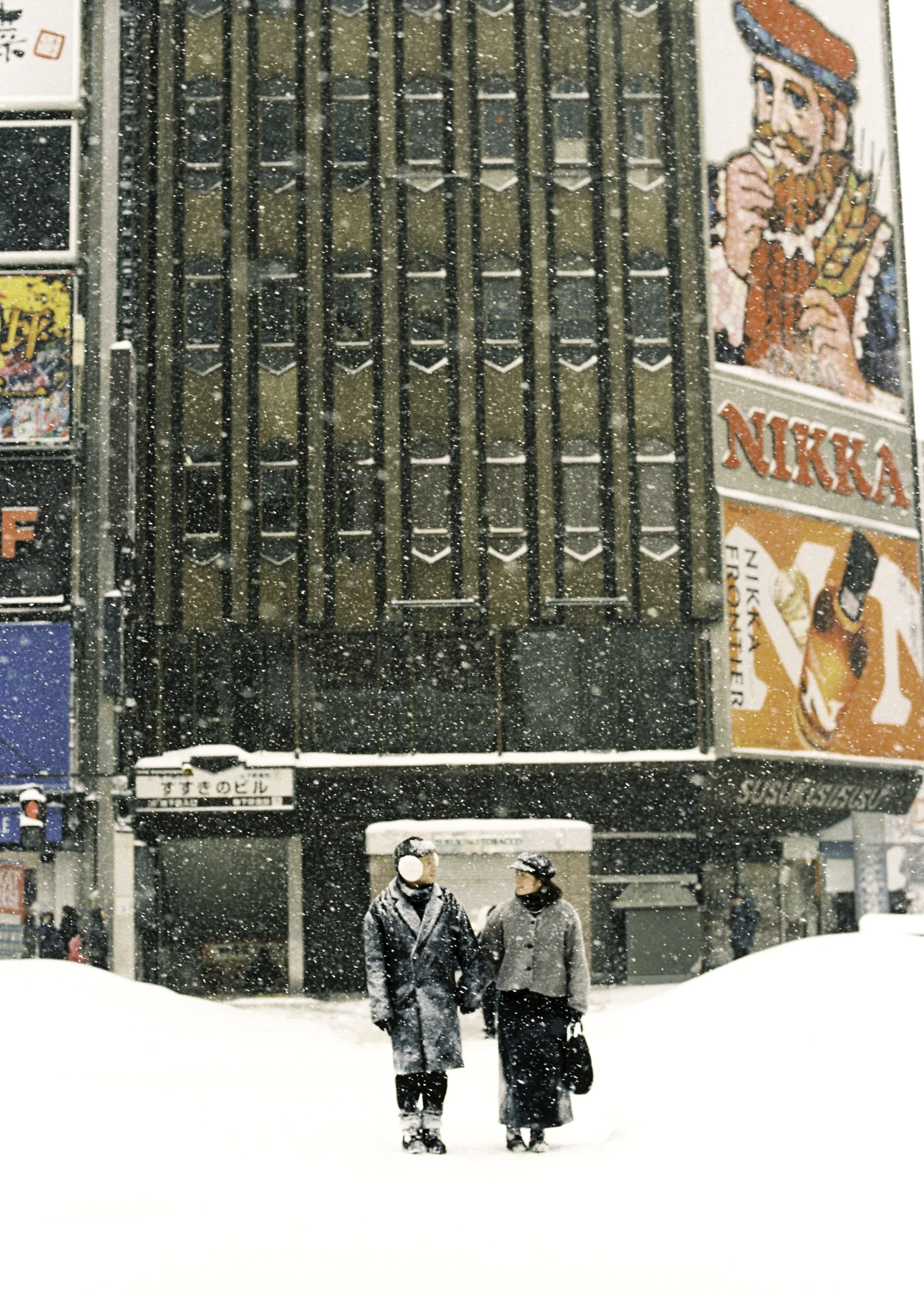 Two people walking in the snow in front of a tall, dark building with billboards, including a large billboard advertising Nikka whiskey, and snow falling heavily. Couple photoshoot in Sapporo.