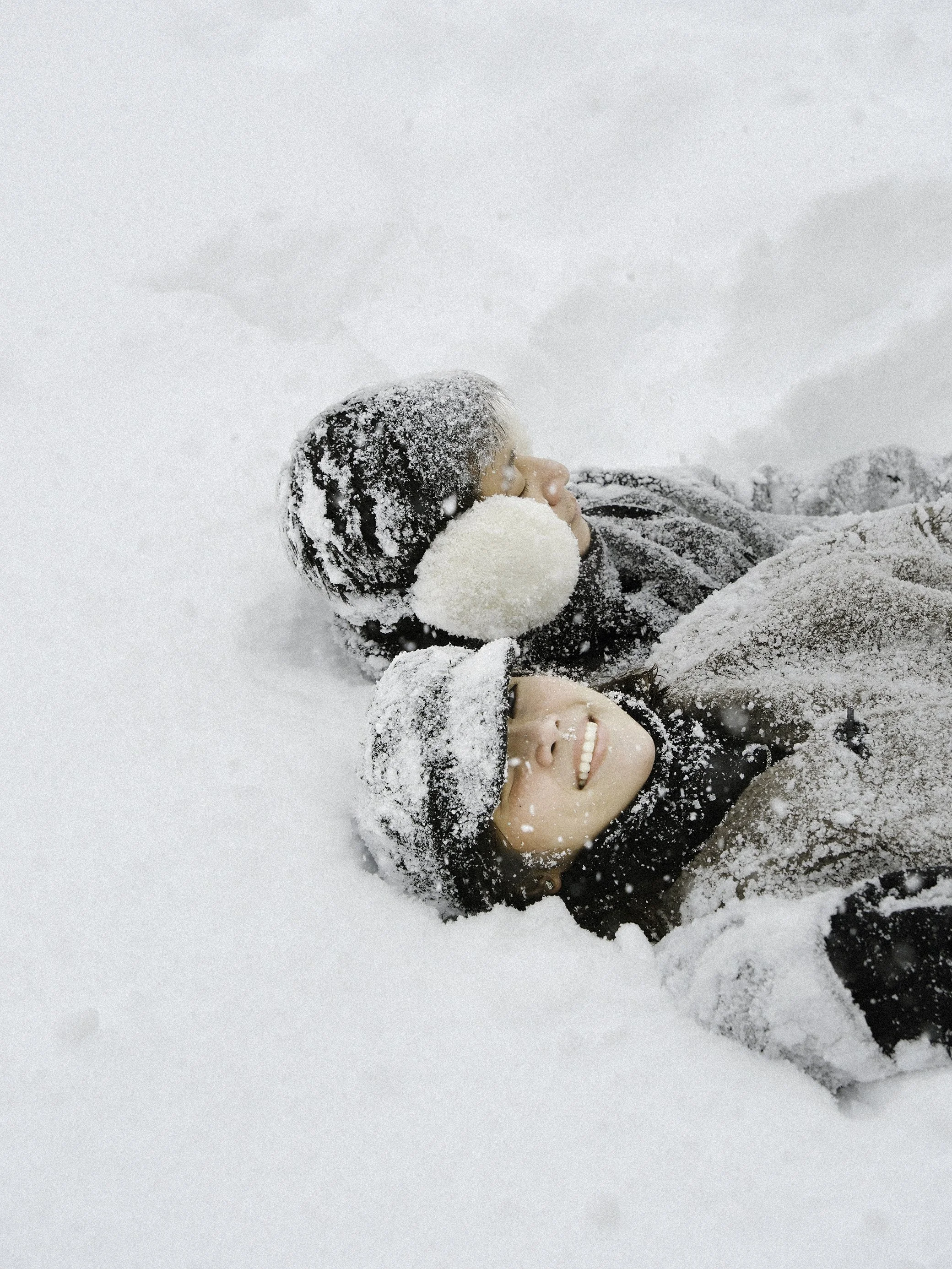Two people lying on snow, smiling and enjoying winter, wearing winter clothes and hats. Couple photoshoot in Sapporo.