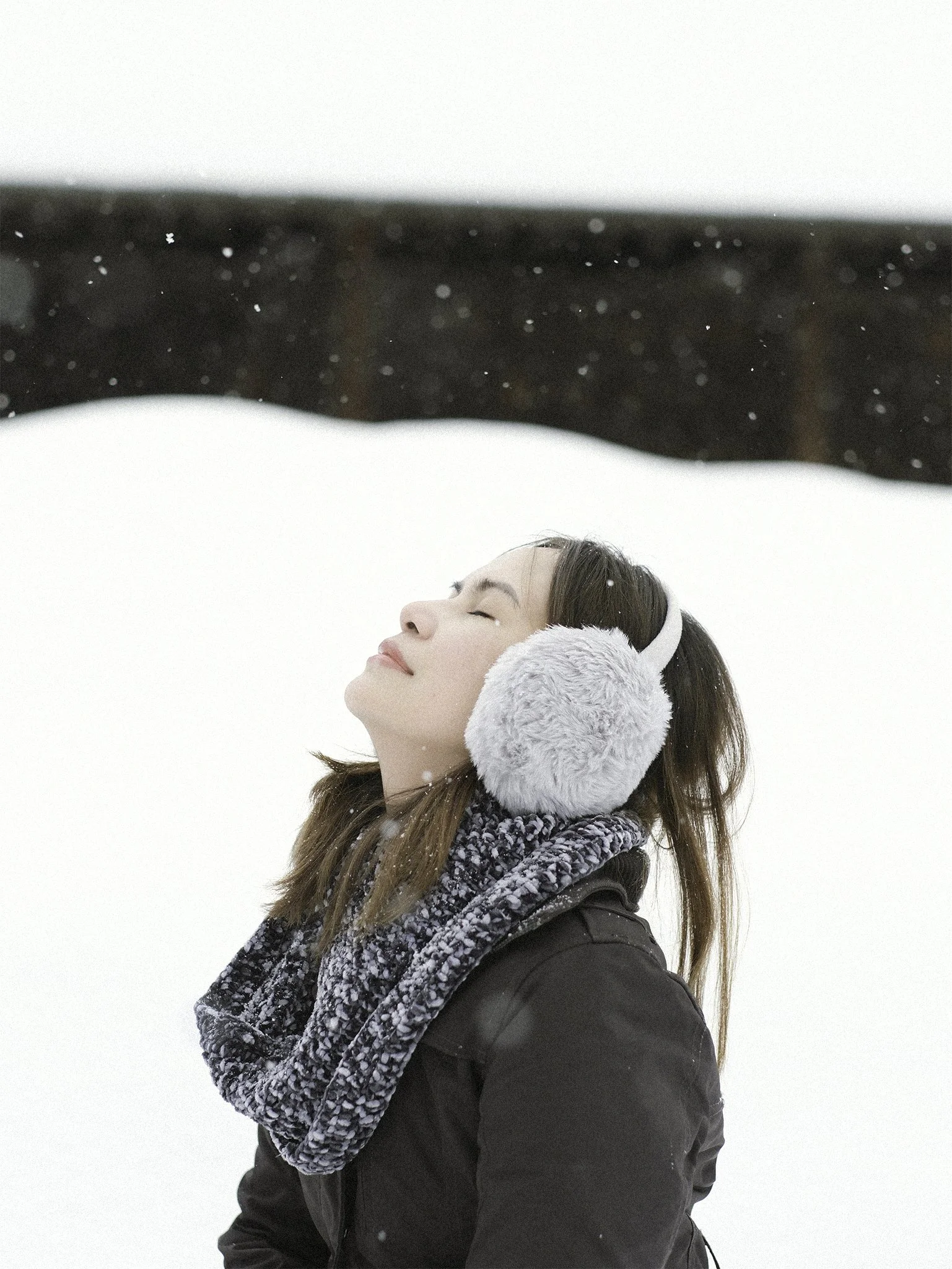 A woman enjoying snow, wearing gray earmuffs, a gray scarf, and a black winter coat, with her eyes closed and face tilted up.