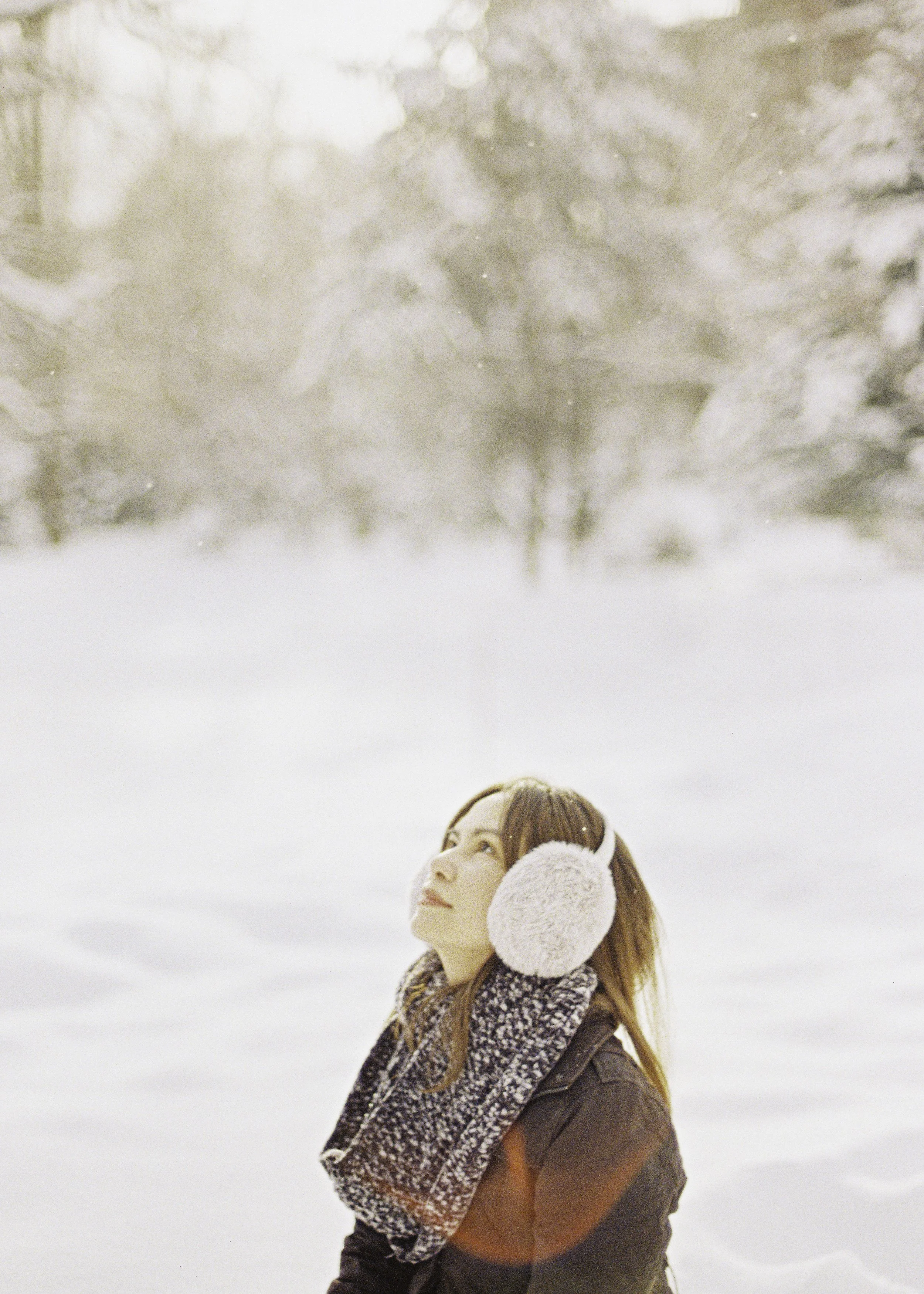 Young woman with long brown hair wearing earmuffs, a scarf, and a jacket standing in snowy landscape, looking upward with snow-covered trees in the background.