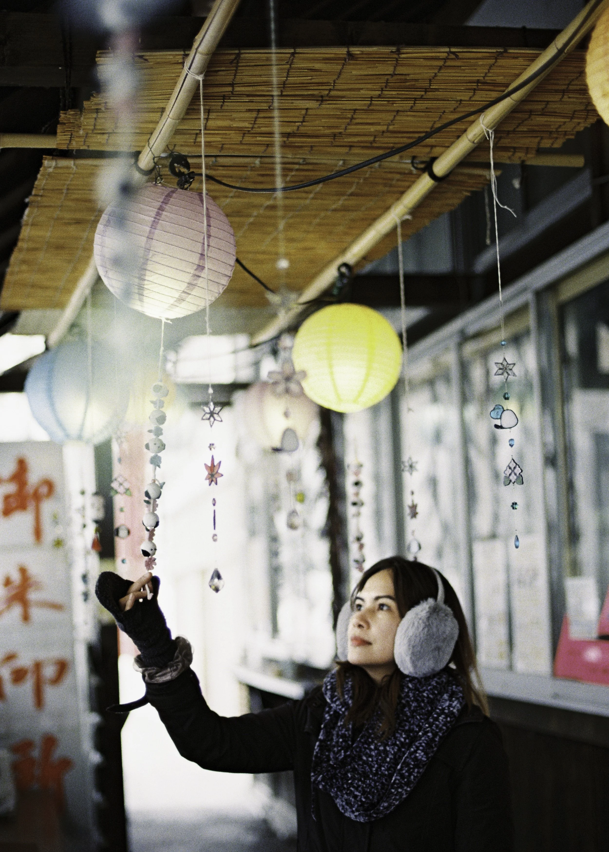 A woman wearing gray earmuffs and a dark coat, holding a string of hanging ornaments, stands under paper lanterns and decorative ornaments hanging from a bamboo and string setup in an outdoor space with storefronts.