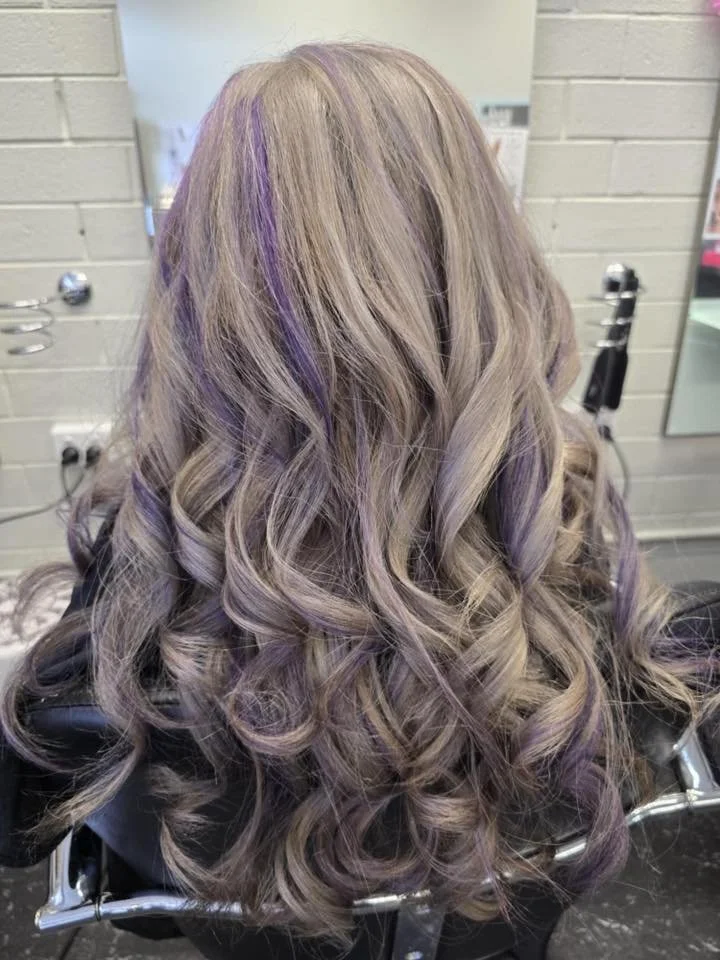 Long, curly hair with blonde and purple highlights dyed in loose curls, over a salon chair in a hair salon.
