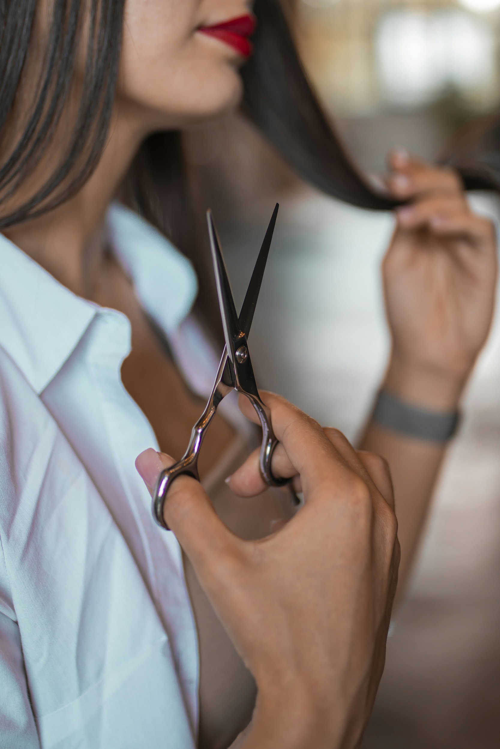 Close-up of a woman with dark hair, red lipstick, and a white top, holding a pair of silver scissors near her hair, preparing to cut it.