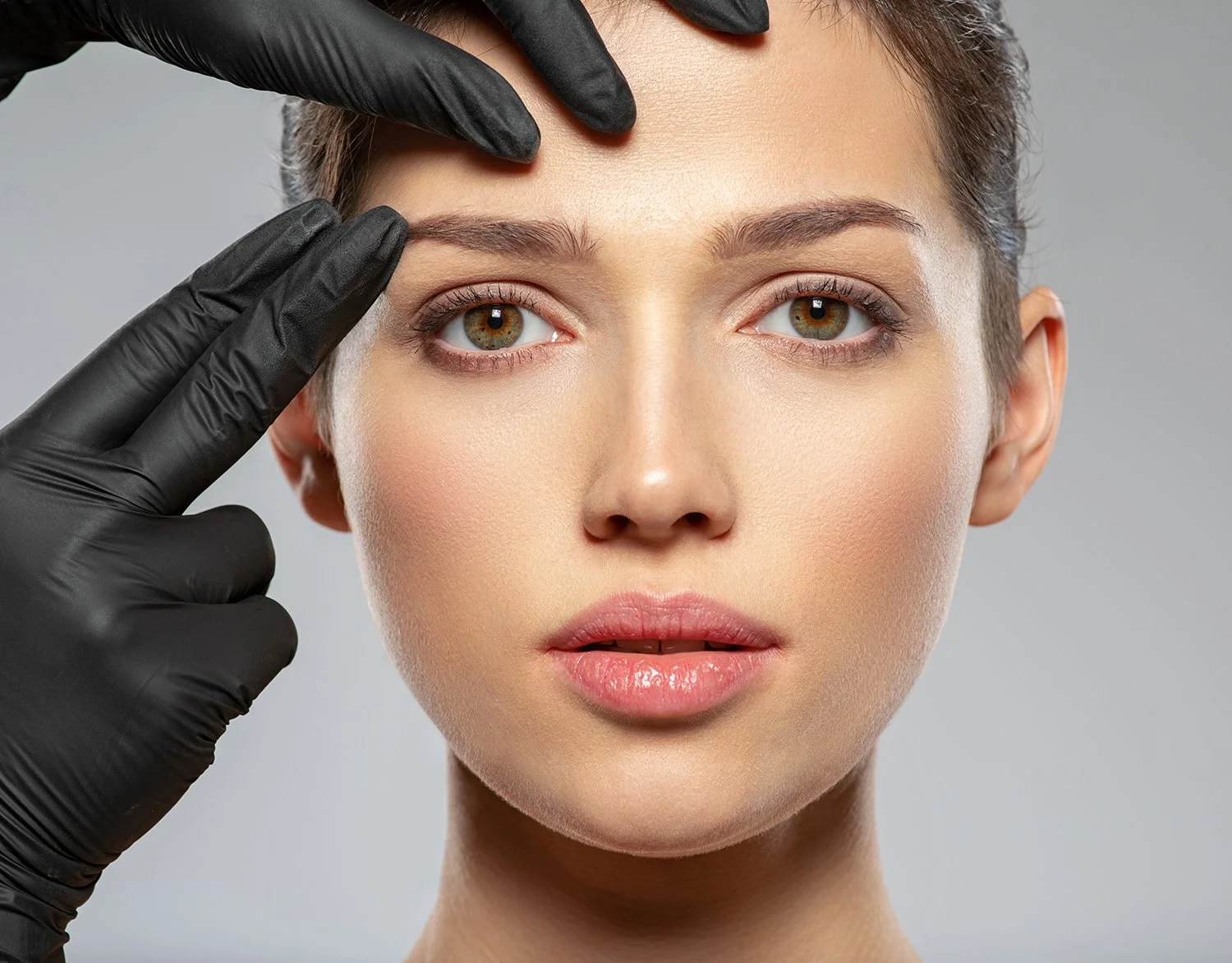Close-up of a woman with clear skin and natural makeup, touching her forehead with gloved hand, in a clinical setting.