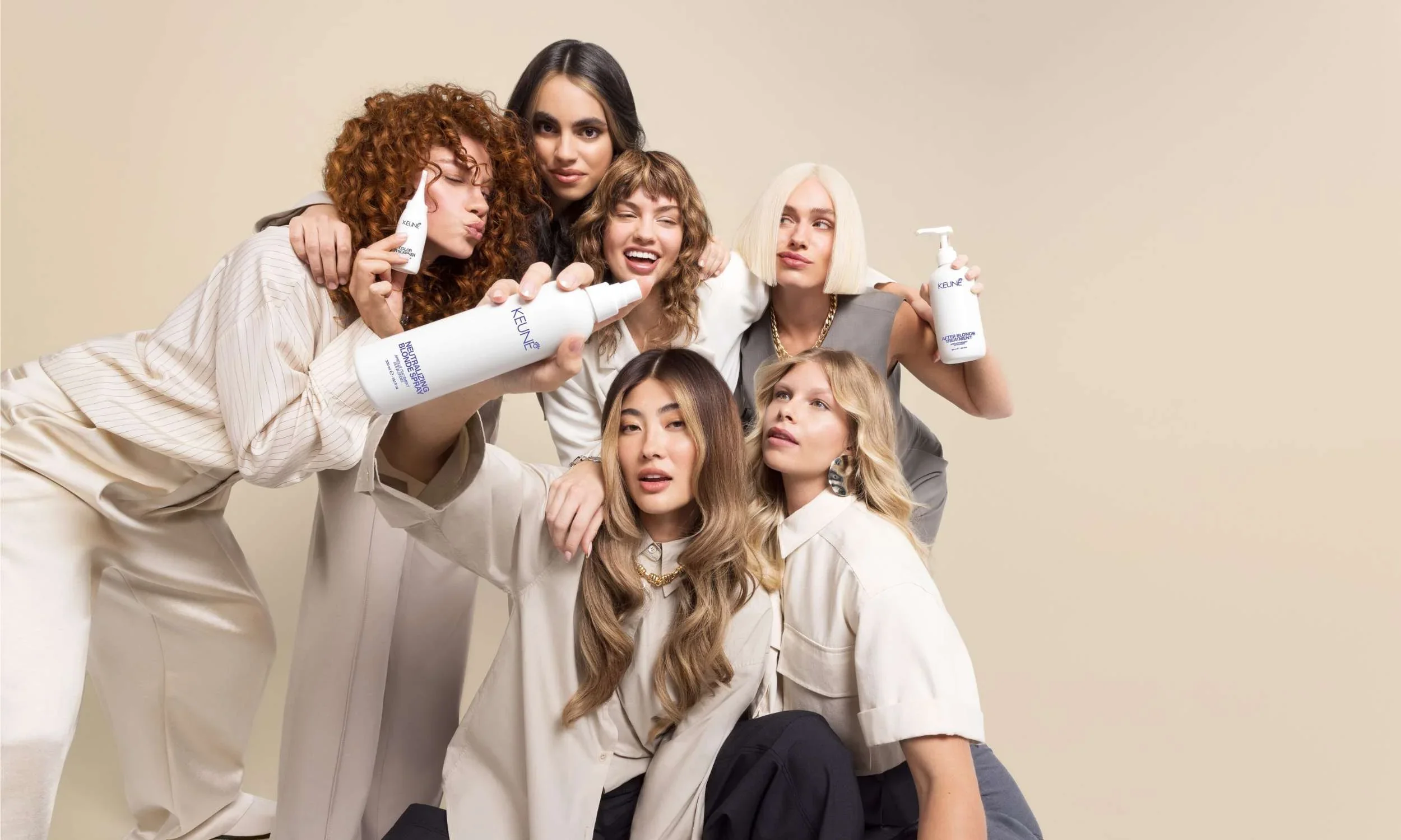 A diverse group of six women with different hairstyles and hair colors, posing together with some holding hair care products, against a plain beige background.