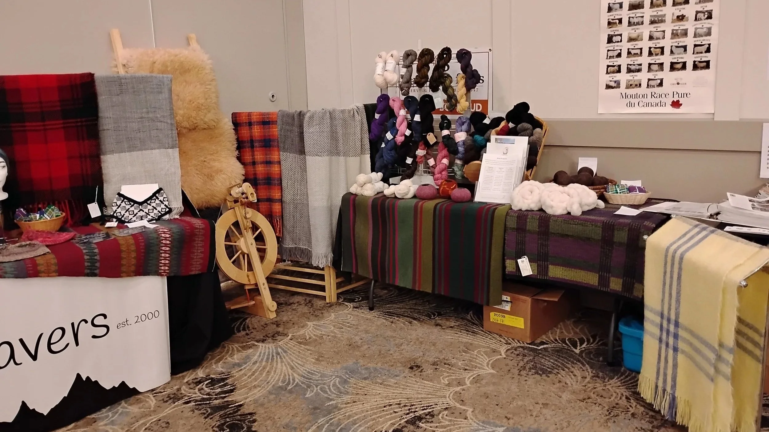 A display at a craft fair featuring colorful woven blankets, fluffy scarves, and yarn skeins, with a spinning wheel in the center.