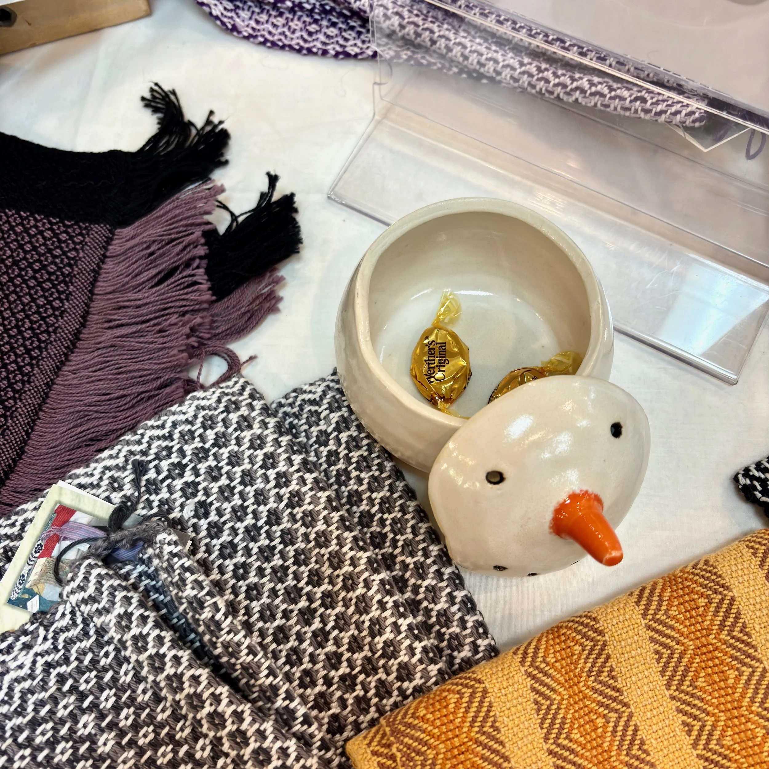Decorative snowman-shaped container with a carrot nose and black eyes, placed on a table with assorted textiles, candies, and a transparent box.