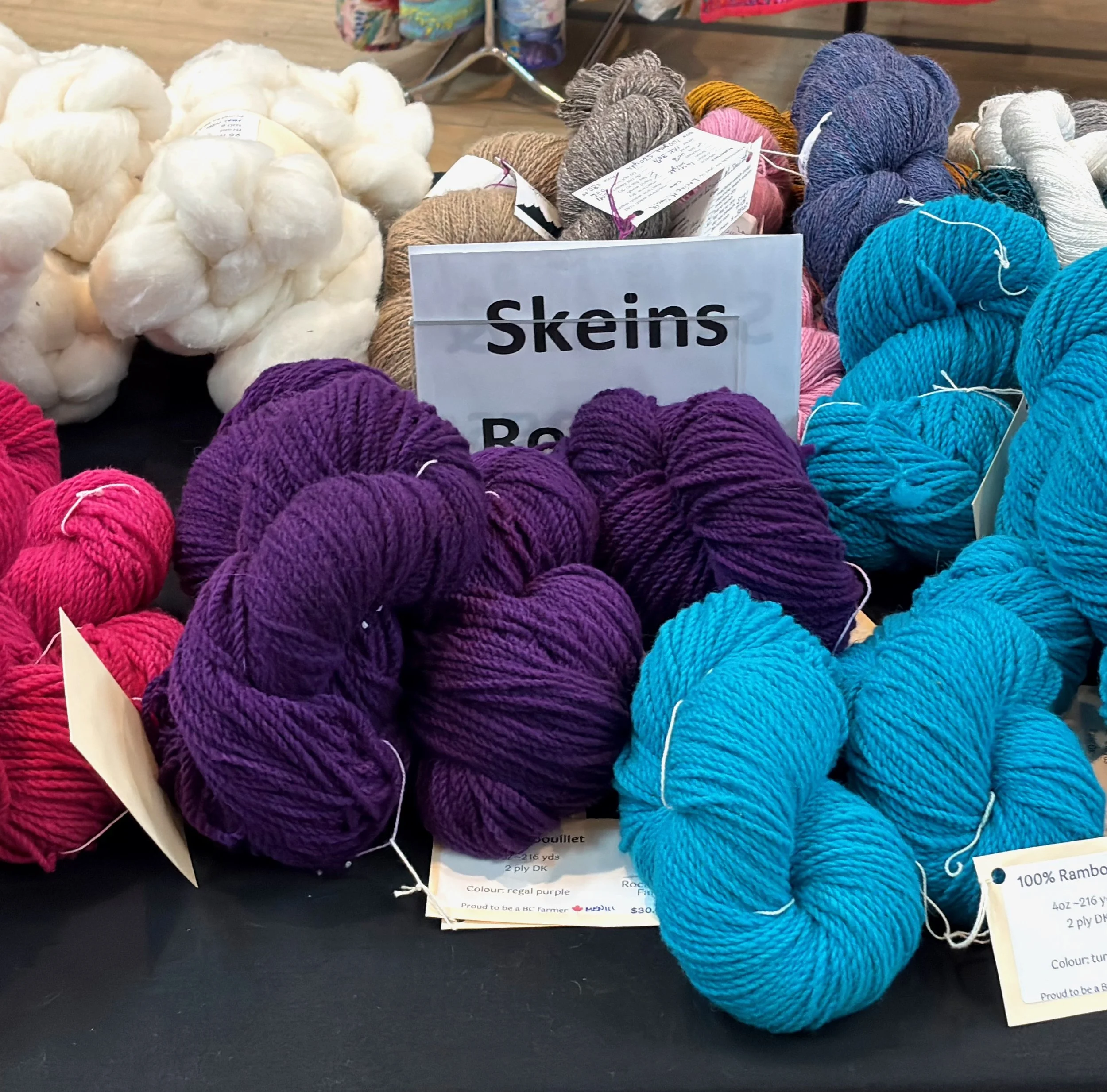 Bundles of colorful yarn on display, including purple, blue, pink, white, and multicolored skeins, with a sign reading 'Skeins'.