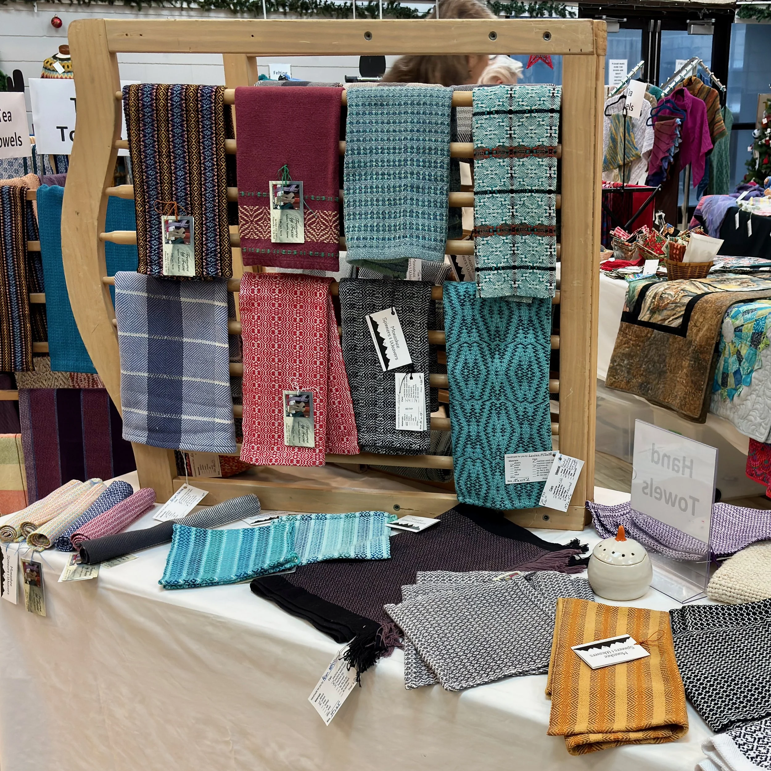 Display of colorful woven scarves and textiles at a craft fair or market, with a wooden rack holding various patterned scarves.