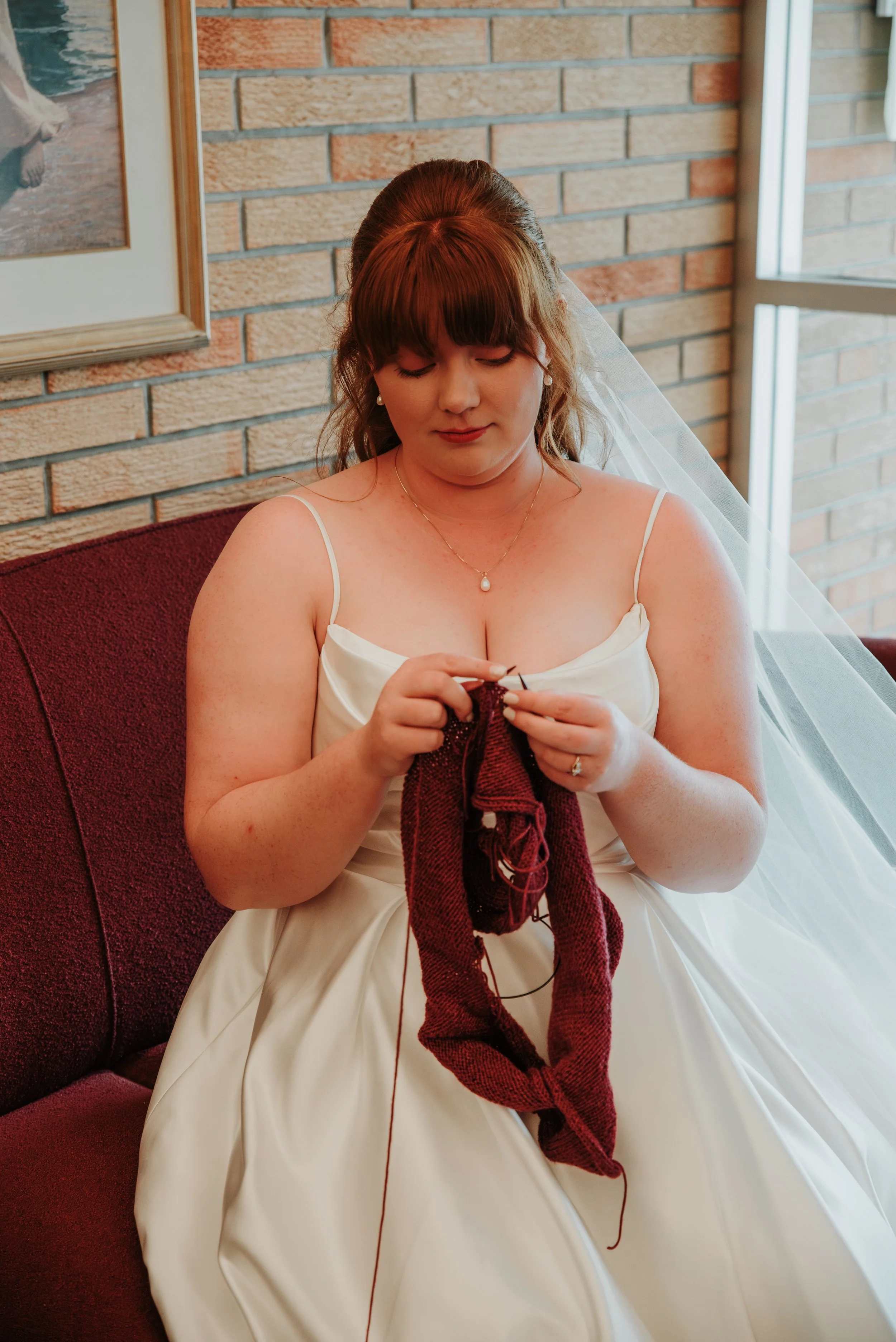 A woman in a white dress sitting on a maroon couch, knitting a red piece of yarn-based fabric, with a brick wall and window in the background.