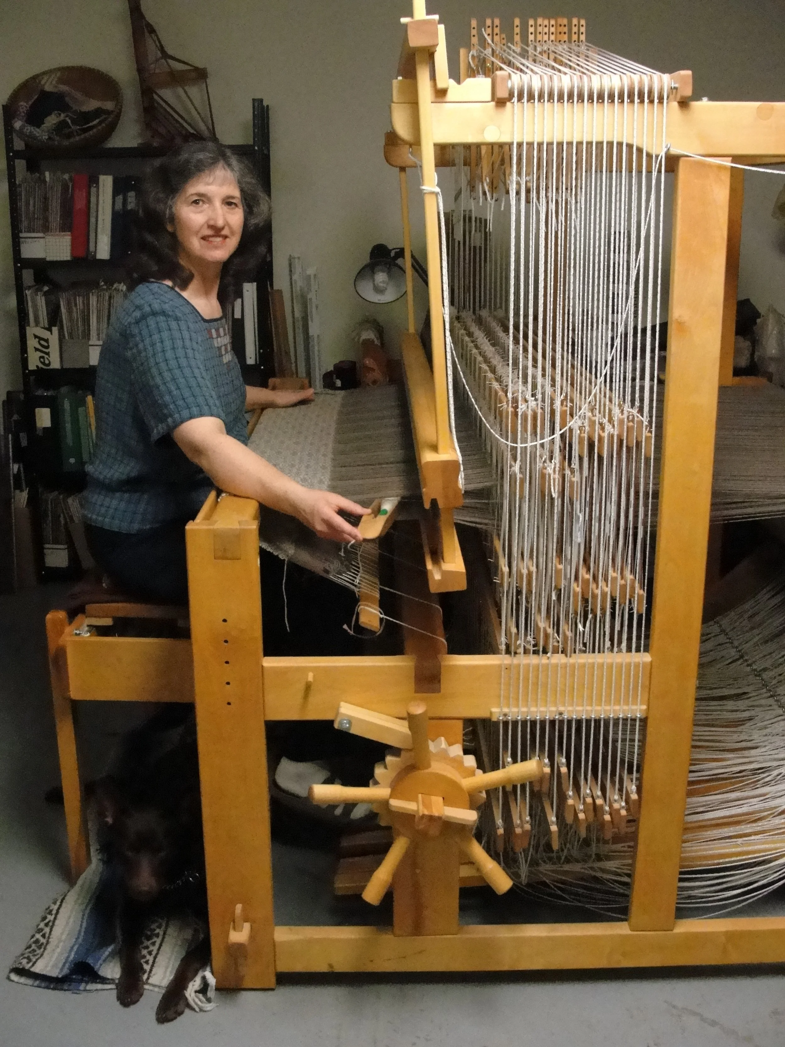 Woman sitting at a floor loom, weaving fabric, with a dog resting underneath the loom.