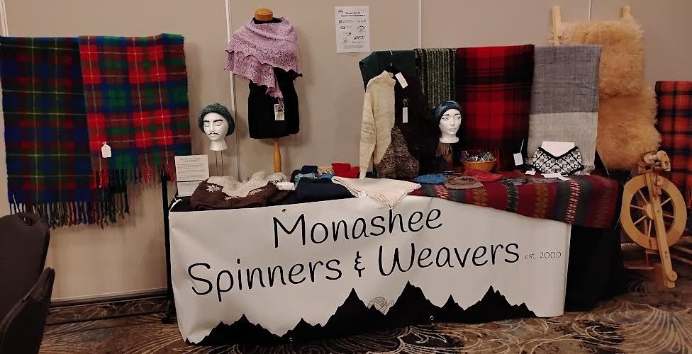 A display booth for Monashee Spinners & Weavers with various scarves, shawls, and fabric samples, made from tartan or plaid patterns, along with weaving equipment. The table has a sign with their name and establishment year, 2000.