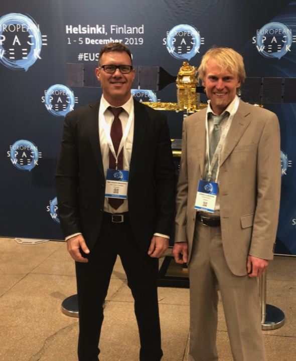 Two men in suits standing in front of a backdrop with the text 'Helsinki, Finland 1-5 December 2019 #EUSEA,' at a European Space Week event, with a model of a satellite in the background.