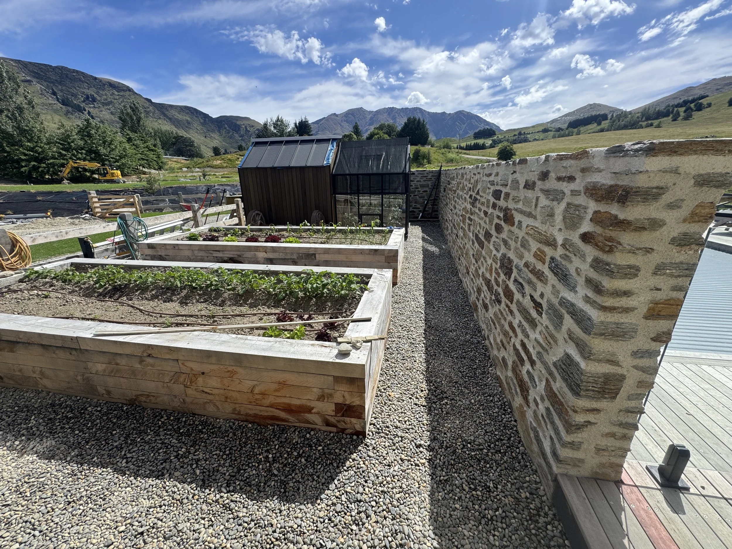 Garden beds with growing vegetables and herbs, stone wall, and mountain landscape in the background on a sunny day.
