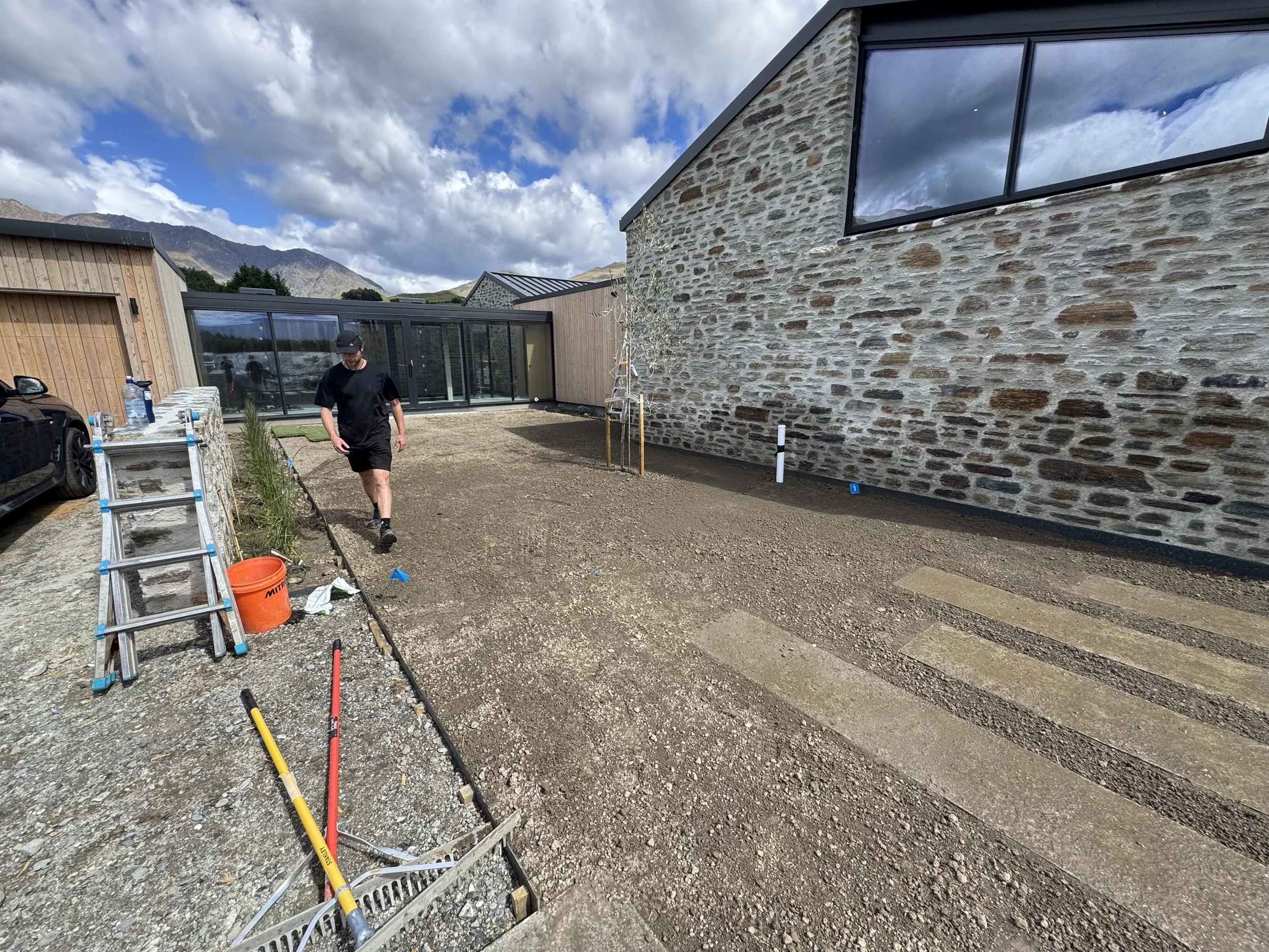 Construction worker walking on gravel driveway with tools and construction materials nearby, adjacent to a modern house with stone and wood exterior.