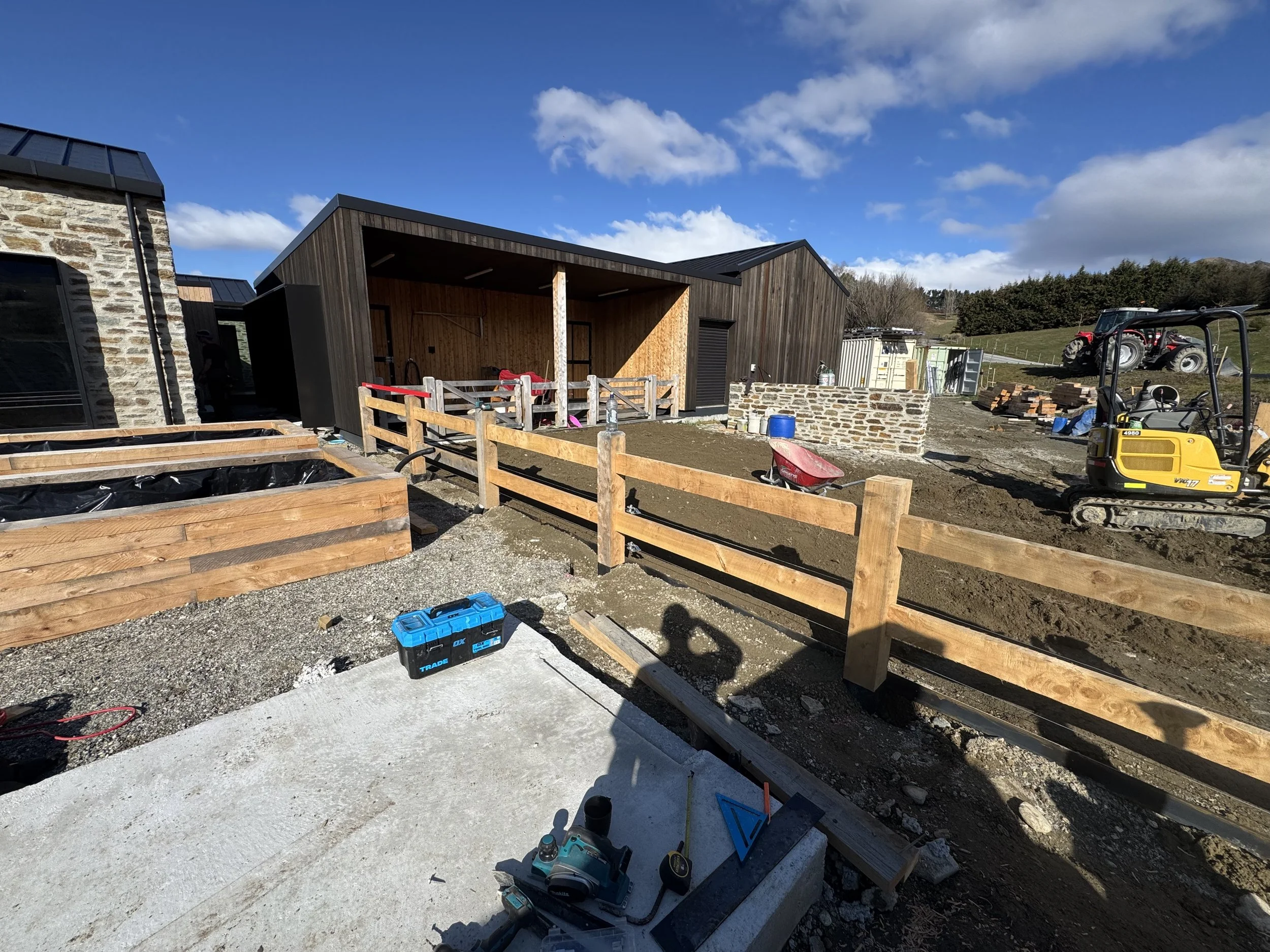 Construction site with wooden fences, tools, and a small building under construction, with a tractor and blue sky in the background.