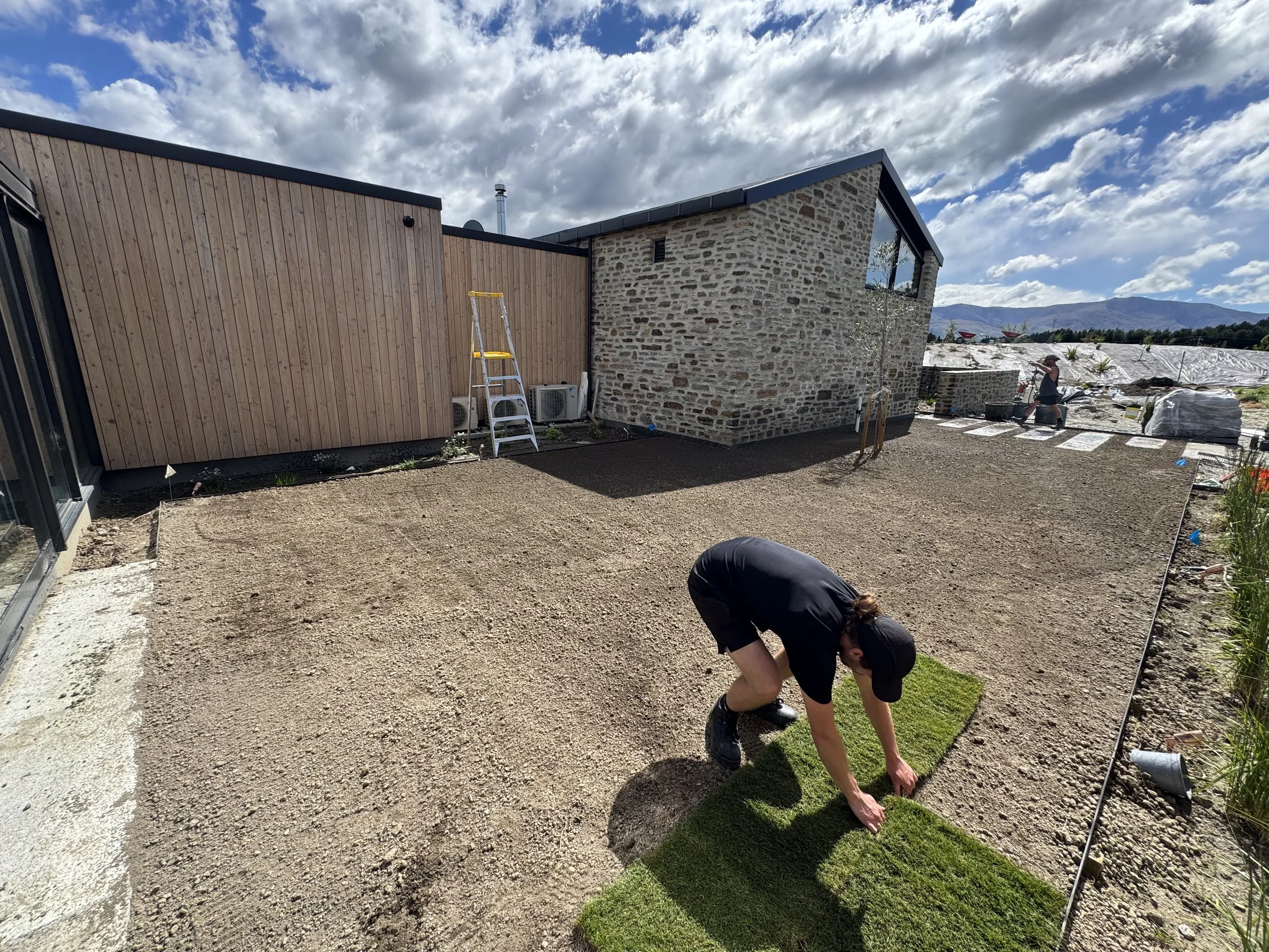 A person laying down sod on a dirt yard outside a house with a stone and wooden exterior on a sunny day.