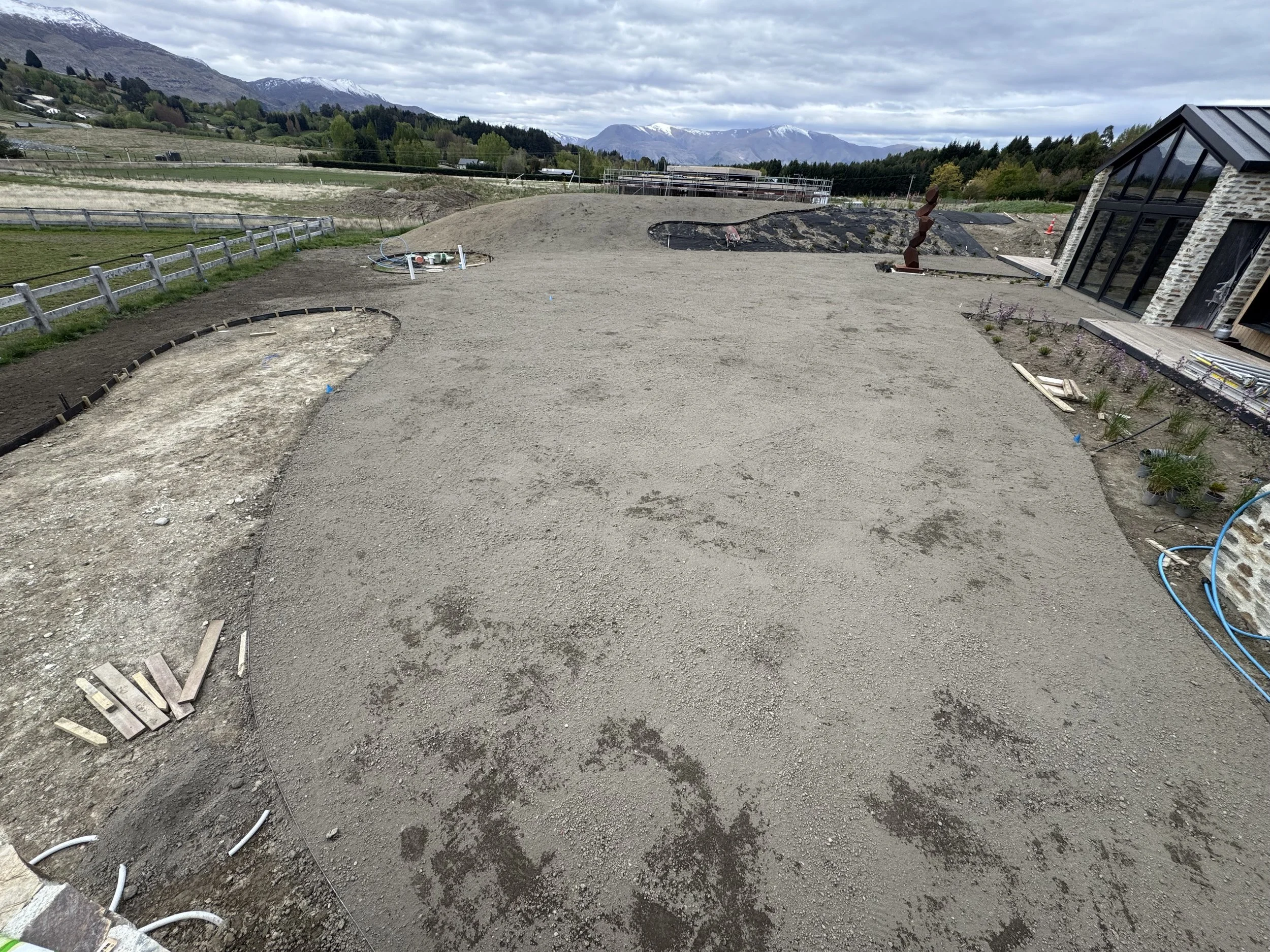 Under construction garden or backyard with a gravel pathway, plants, and a modern house with large glass windows, surrounded by mountainous landscape.