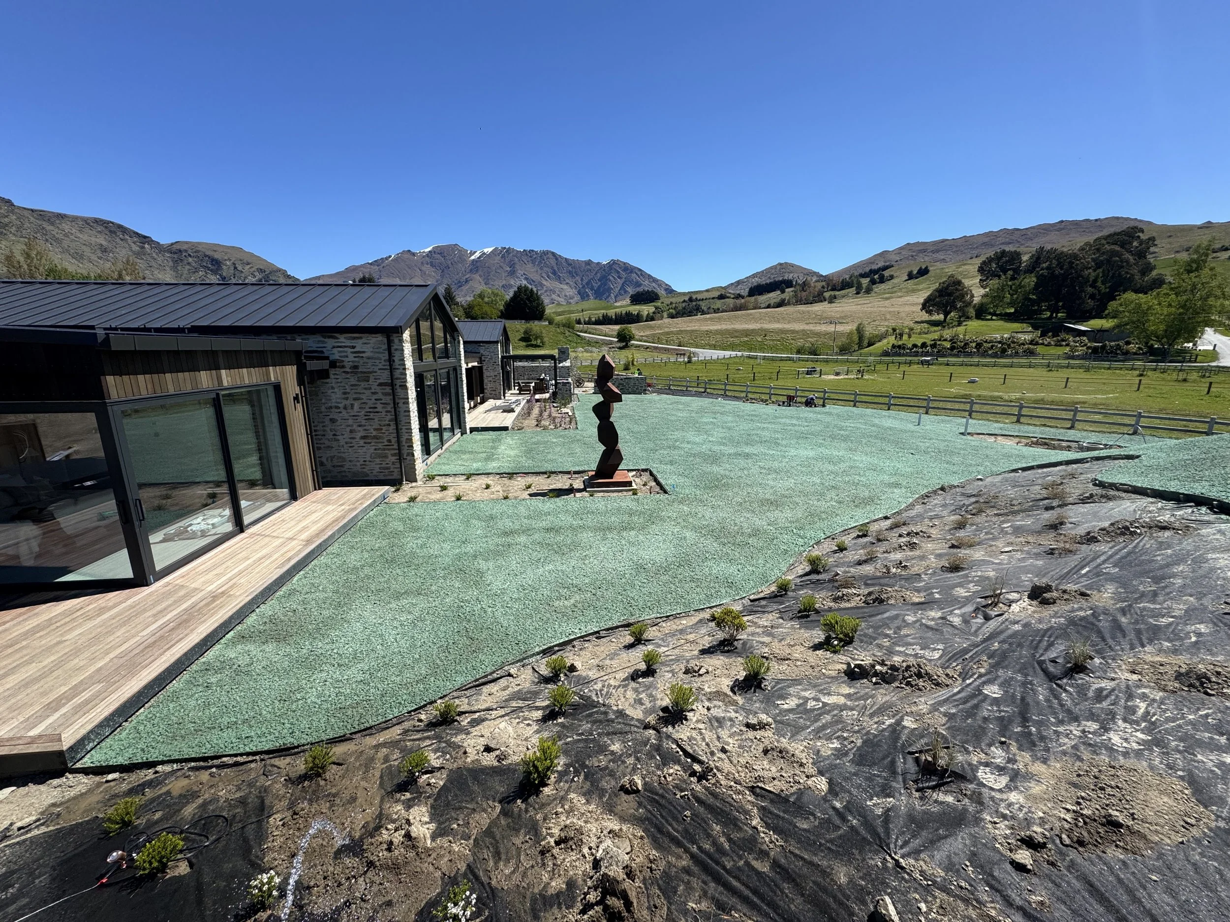 Modern house with large glass windows, outdoor deck, and landscaped yard on a hillside with mountains in the background under a clear blue sky.