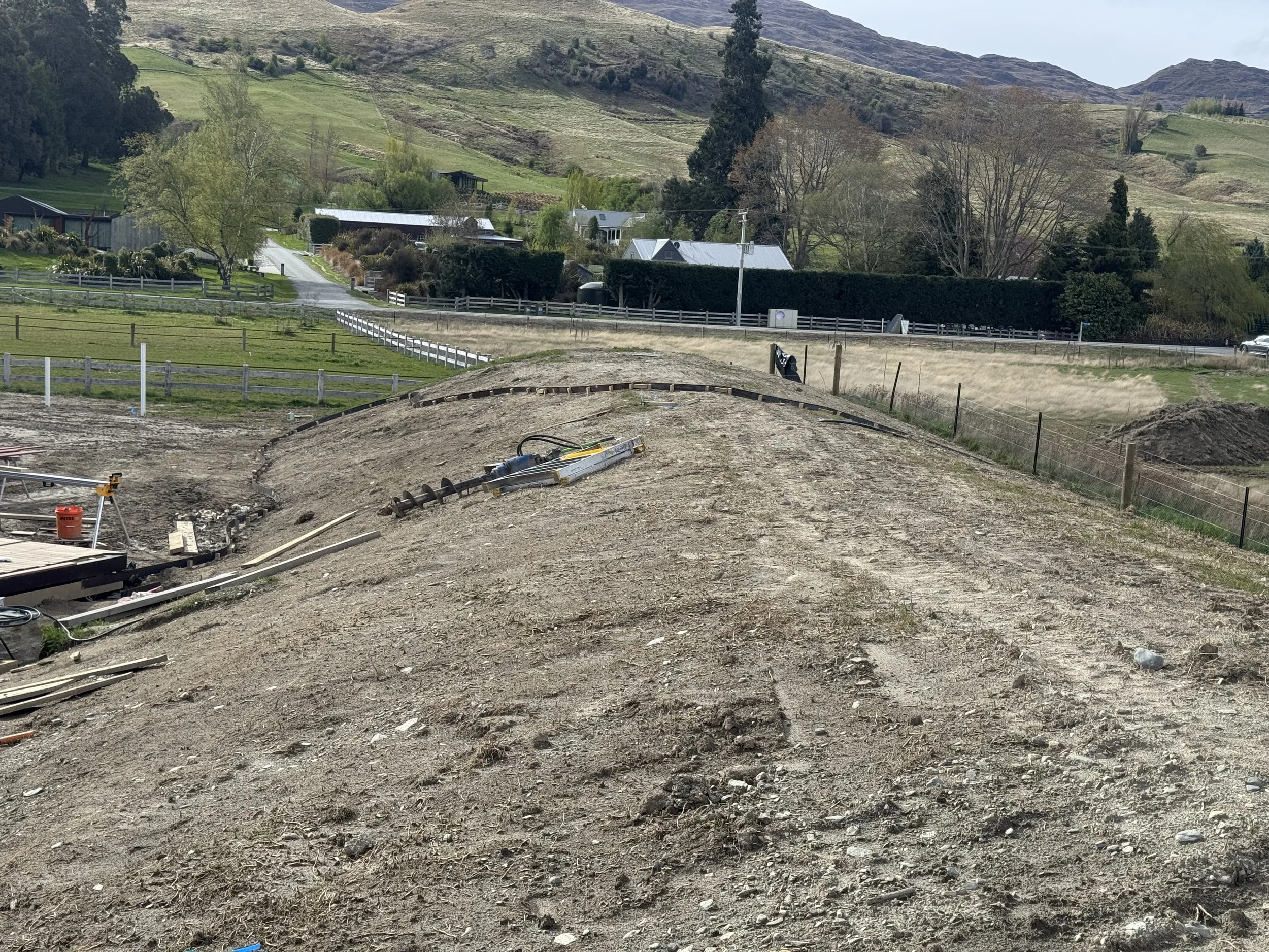 Construction site on a hill with dirt and building materials, bordered by a fence, with green fields, trees, and hills in the background.
