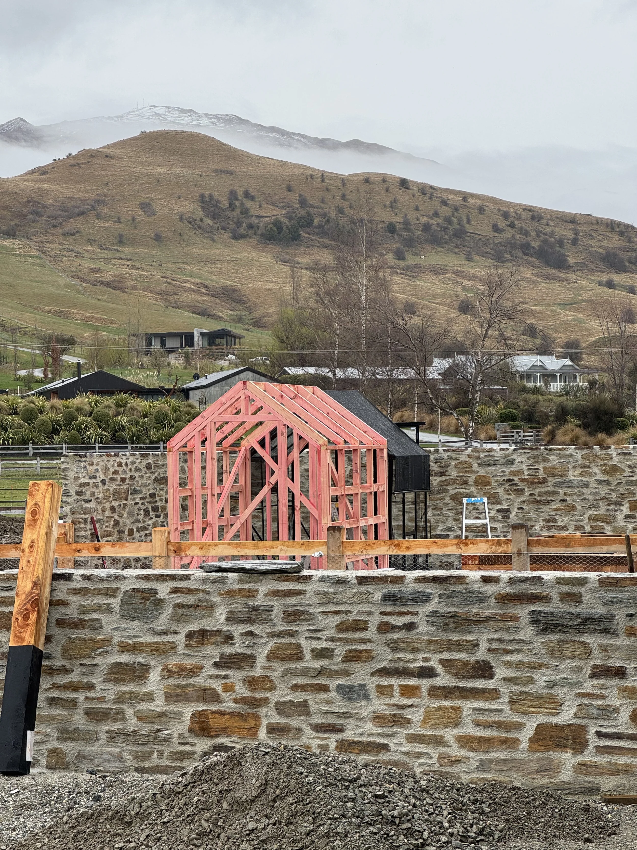Construction site with a stone wall, wood framing for a structure, and a ladder in front of a scenic mountain landscape with cloudy skies.