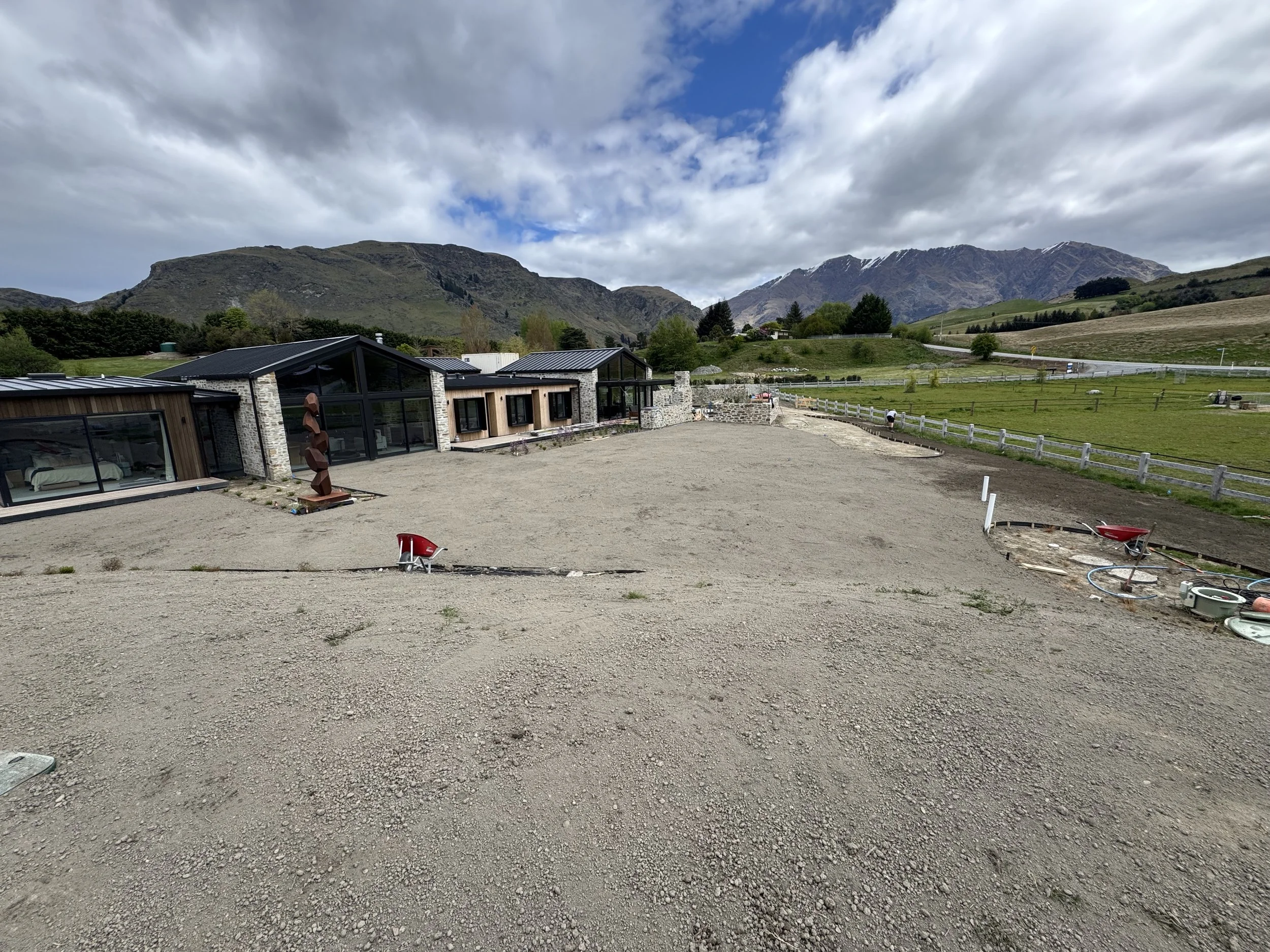 Under construction area with gravel ground, some building structures with large glass windows, surrounding green hills and mountains under cloudy sky.