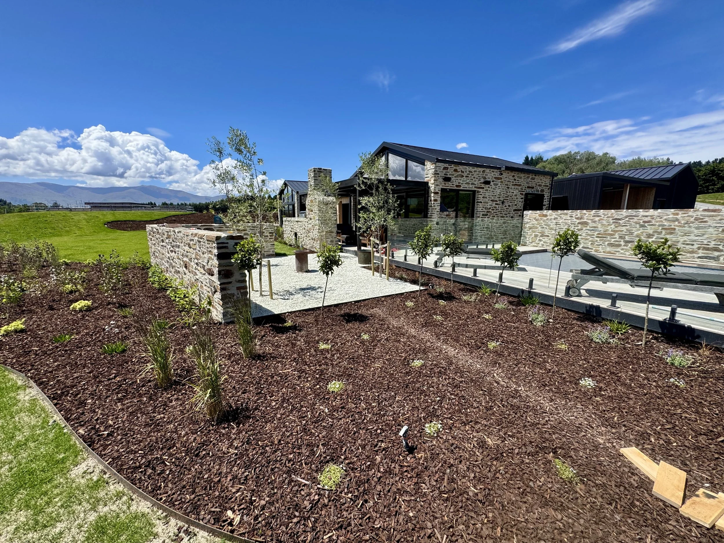 Modern house with stone and black exterior walls, surrounded by landscaped garden with young trees, patio area with benches, under a blue sky with clouds, in a rural setting with mountains in the background.