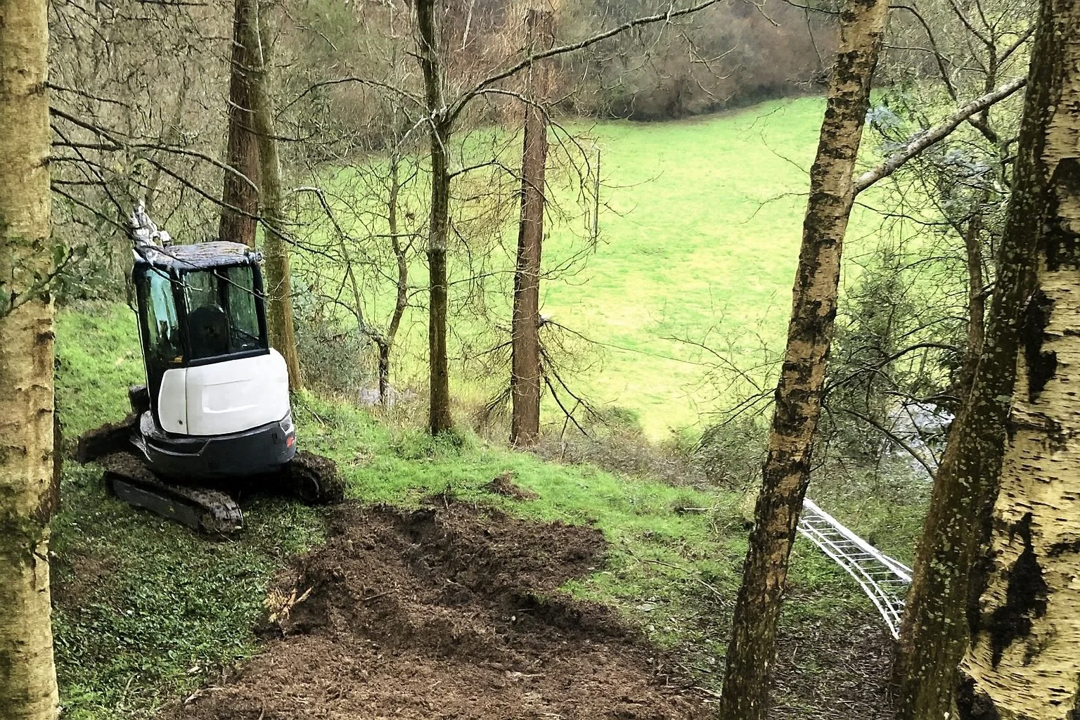A small excavator parked on a muddy patch in a forested area with green grass and trees.