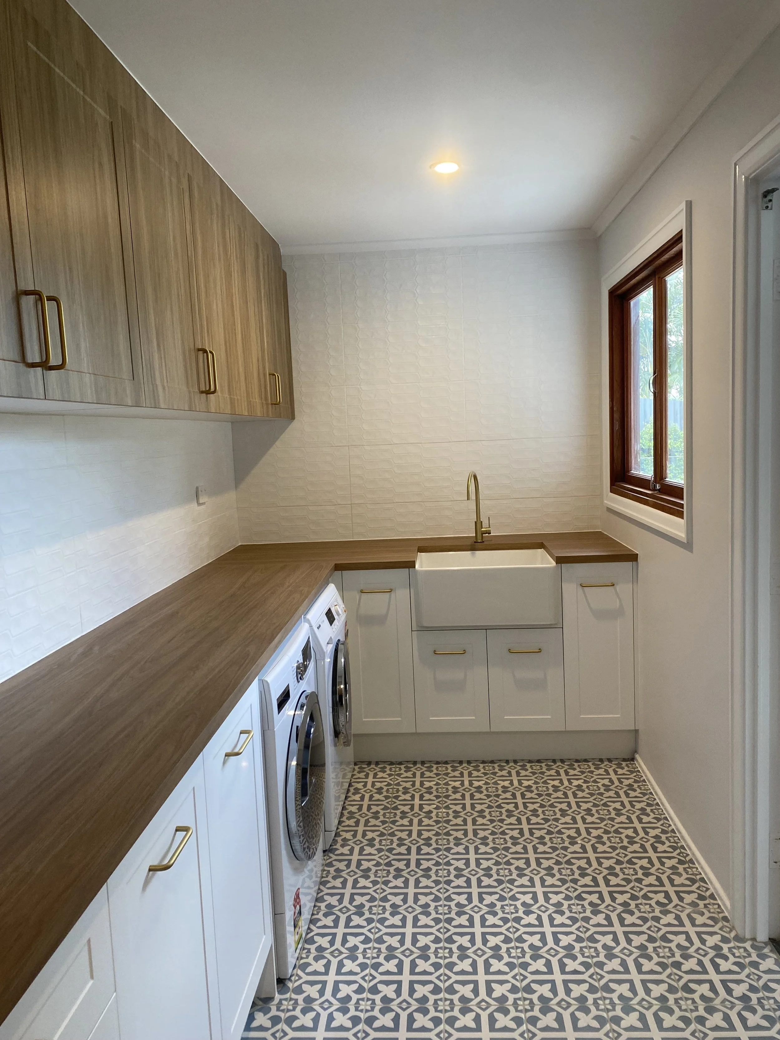Modern laundry room with patterned floor tiles, white cabinets, wood countertops, and a farmhouse sink beneath a window.