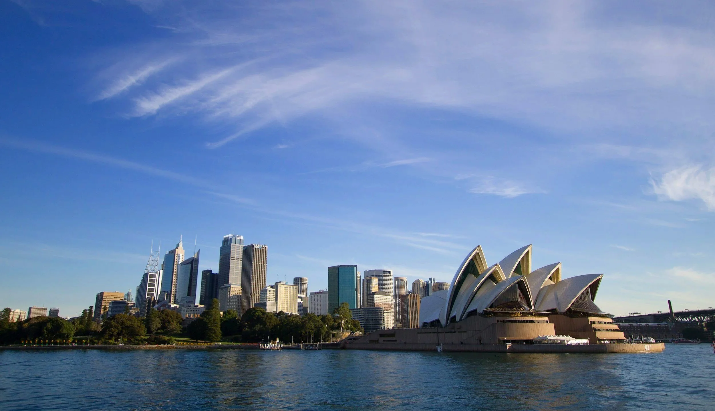 Vista de la ciudad de Sídney con los edificios del horizonte y la Ópera de Sídney en primer plano, junto al río, durante el día con cielo despejado.