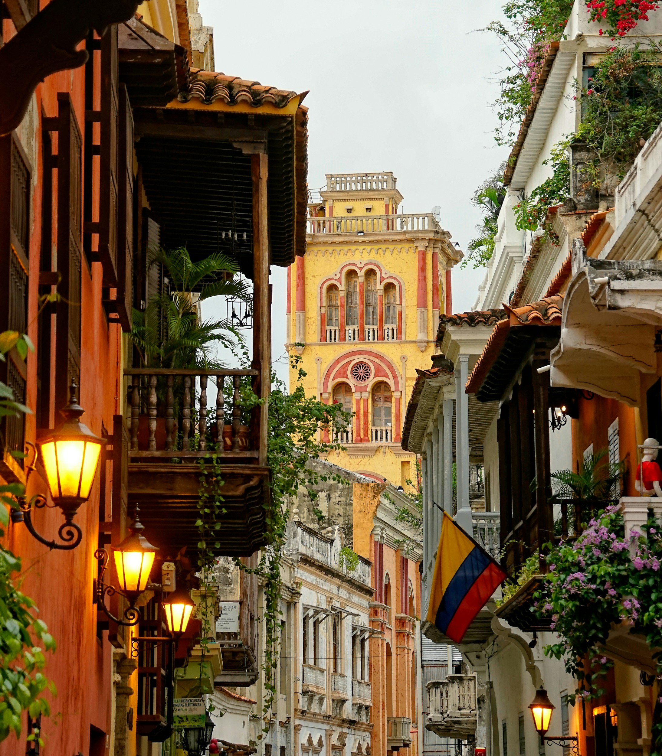 Vista de una calle estrecha con edificios coloniales y un edificio amarillo con detalles en rojo al fondo, en una ciudad colonial, posiblemente en Cartagena, Colombia.