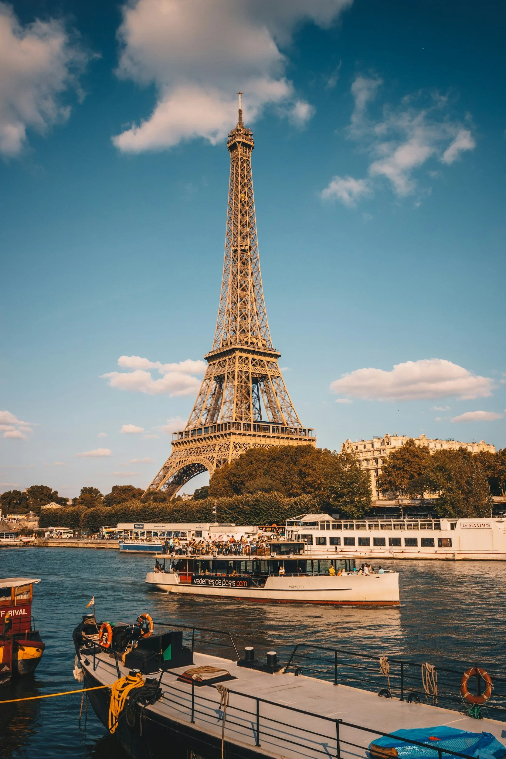Torres en París vista desde el río Sena con barcos y árboles en primer plano y cielo con nubes.