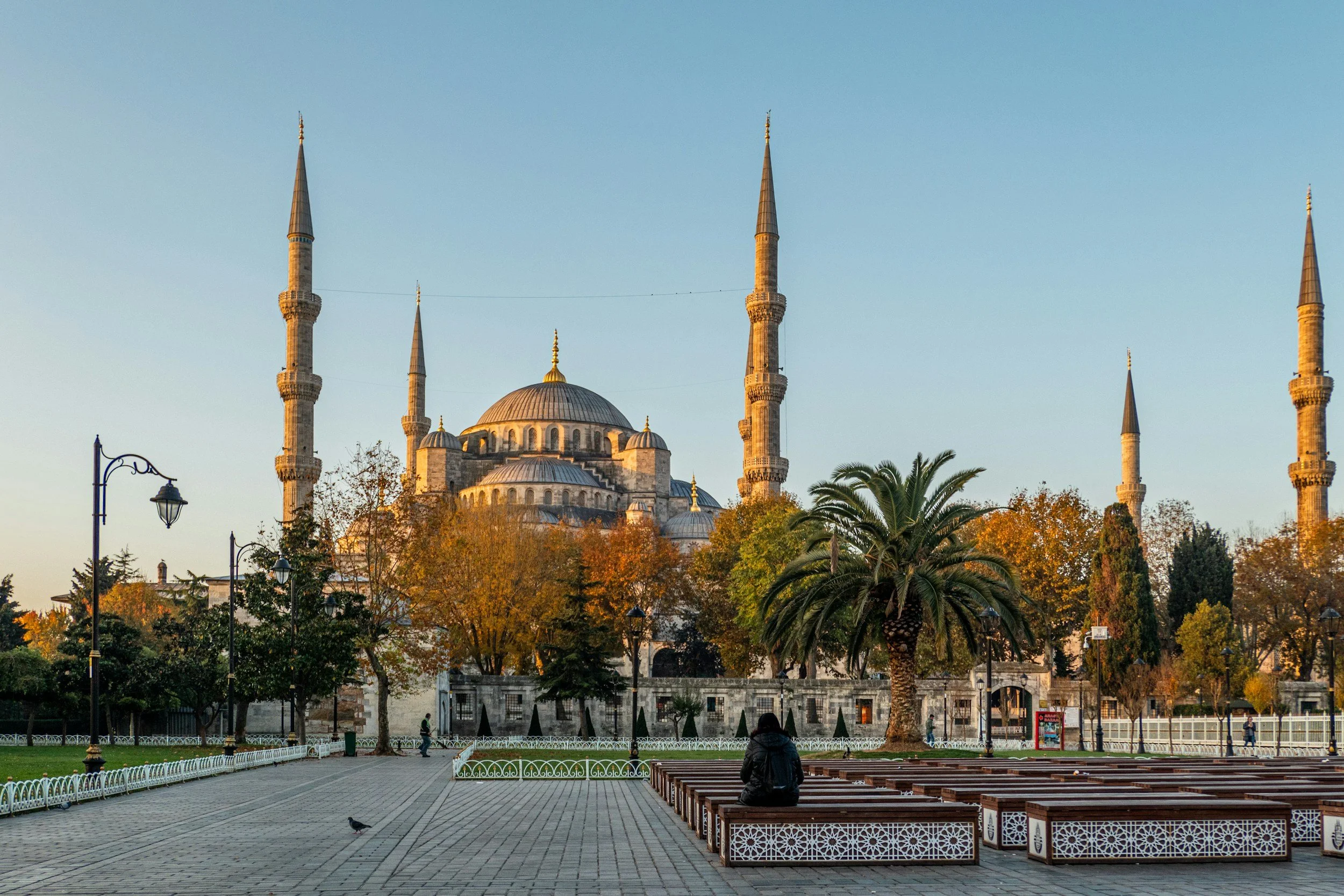 Vista de la Mezquita Azul en Estambul, con árboles en otoño, un sendero peatonal, bancos de madera y una persona sentada en uno de ellos.