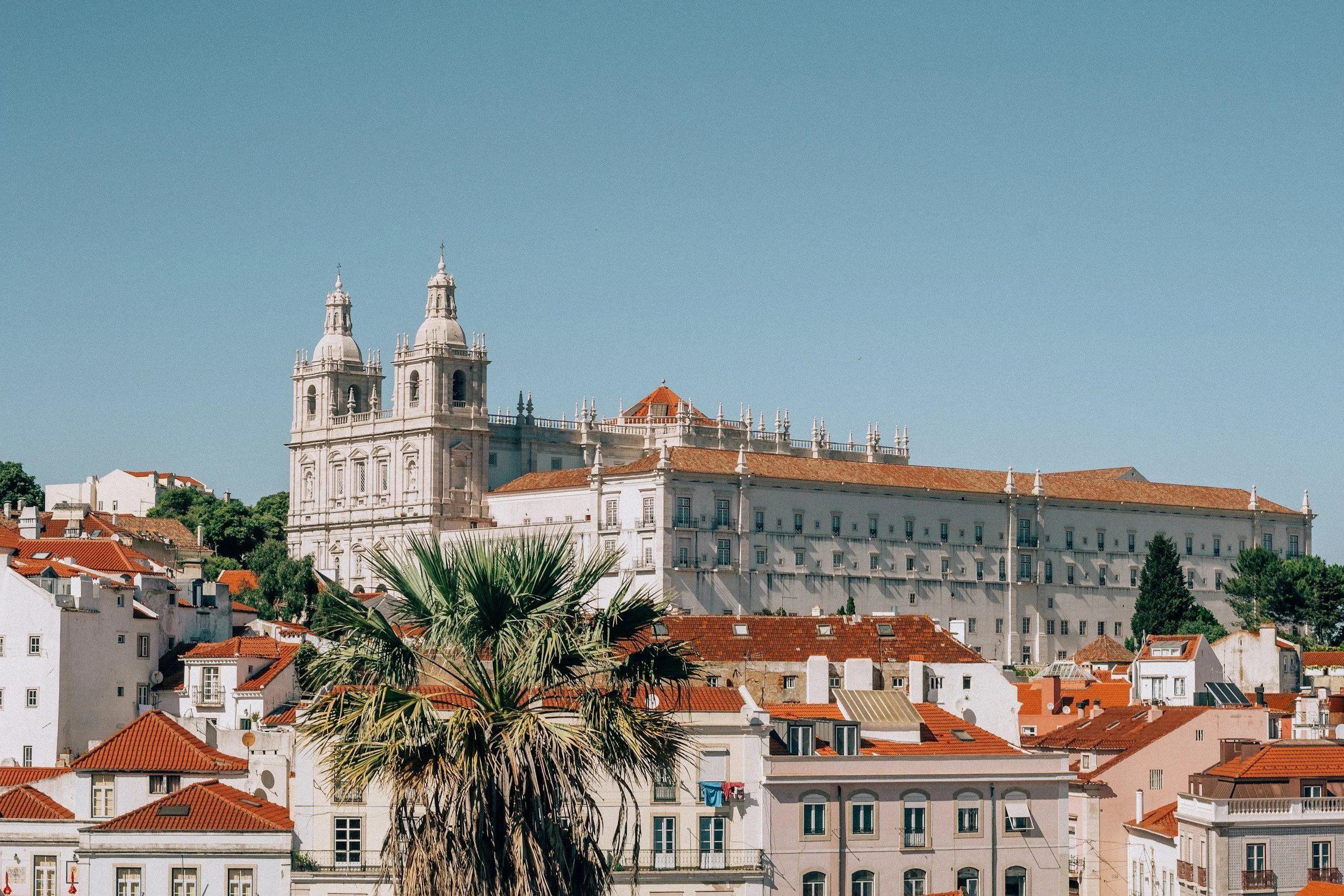 Vista de una ciudad con casas blancas de techos de teja roja y una iglesia grande con dos torres altas y una cúpula, rodeadas de árboles y cielo despejado
