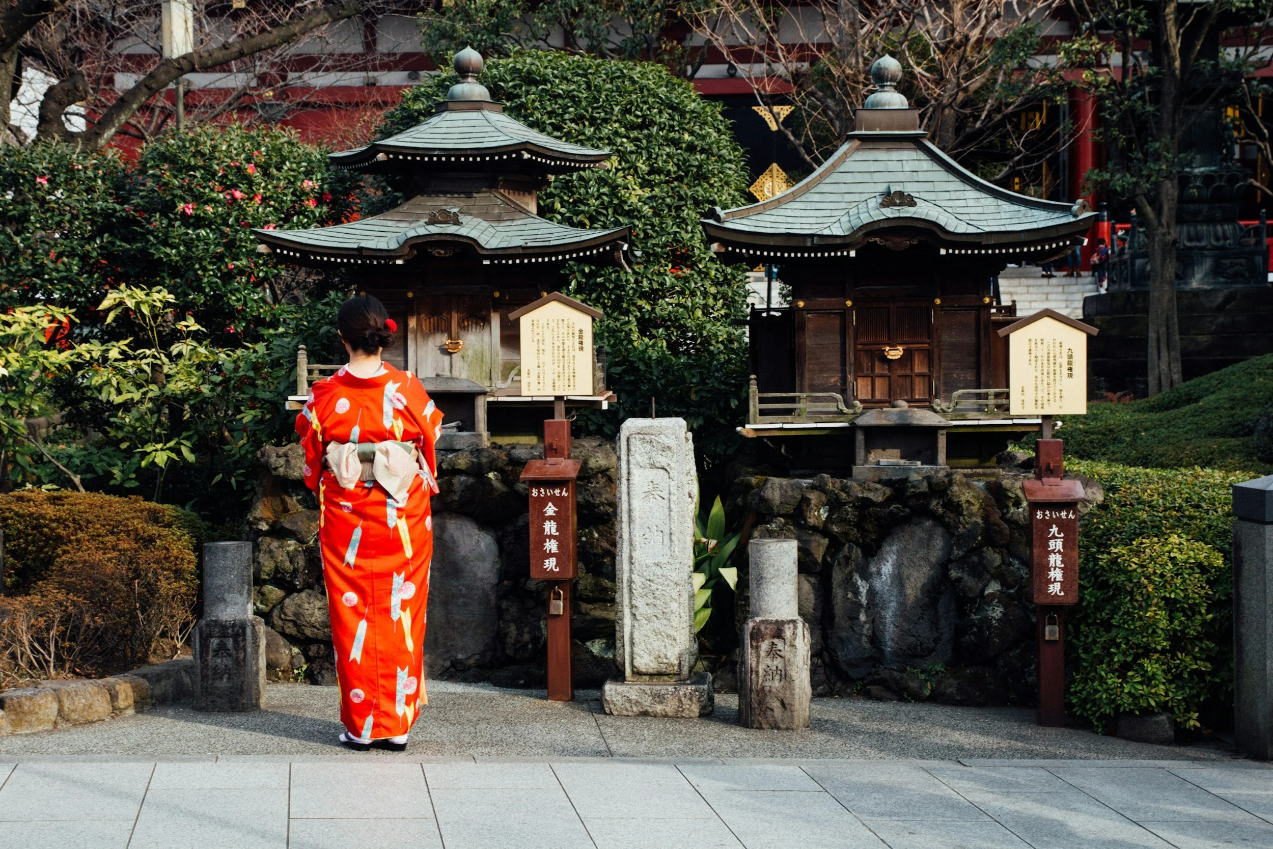 Mujer vestida con kimono rojo observando dos pequeñas estructuras tradicionales de Japón en un jardín.