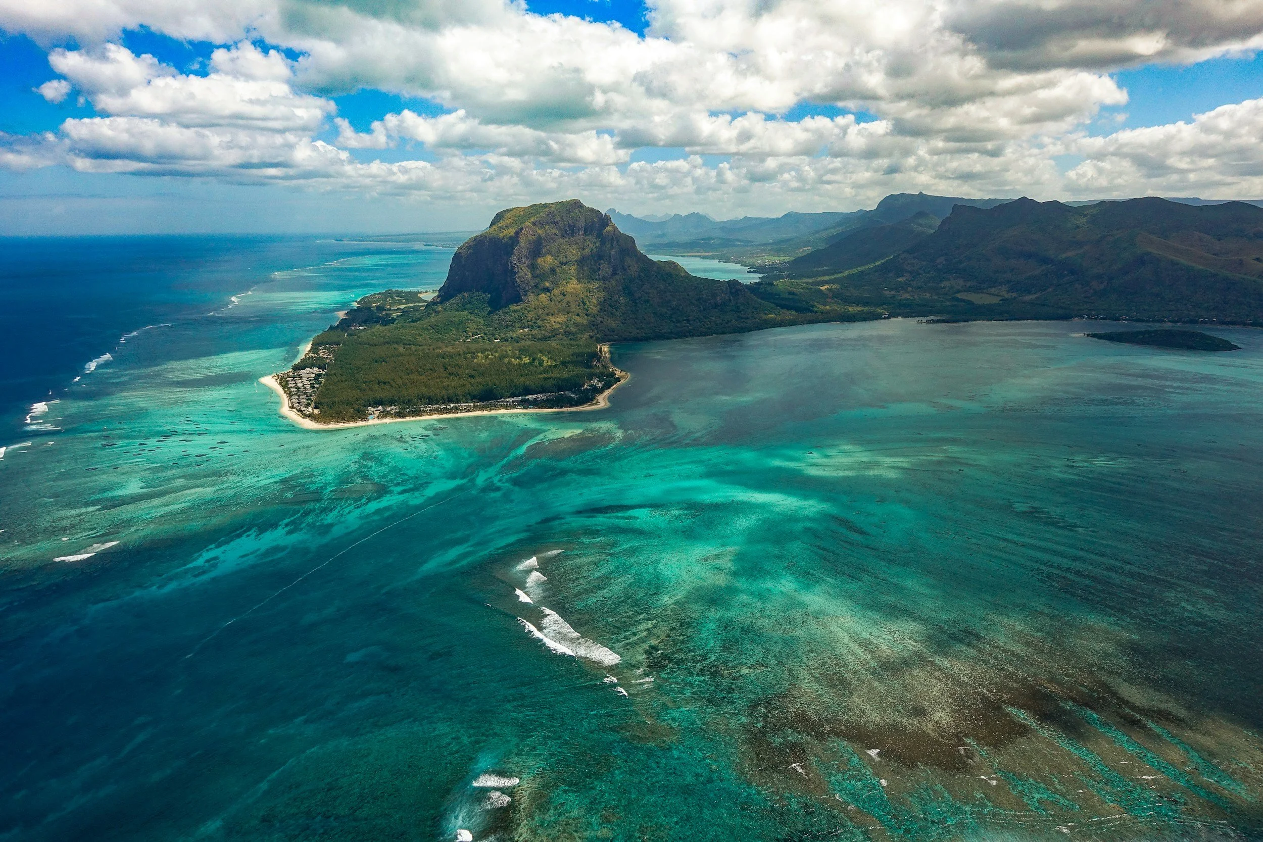Vista aérea de una isla con una montaña alta y áreas de playa y agua turquesa, rodeada por un océano y montañas en el fondo, con cielo parcialmente nublado.