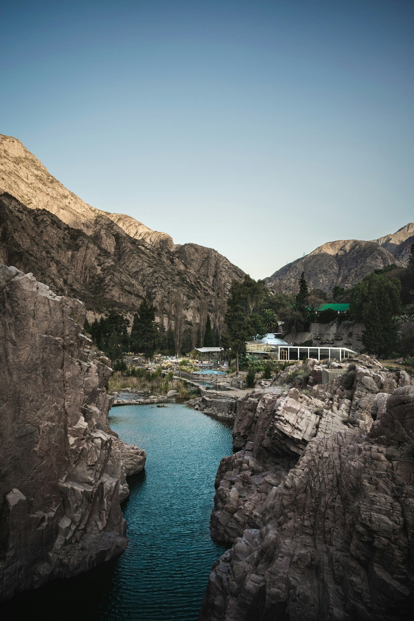 A serene canyon with steep rocky walls on either side and a calm blue river flowing through it, surrounded by trees and mountains under a clear sky.