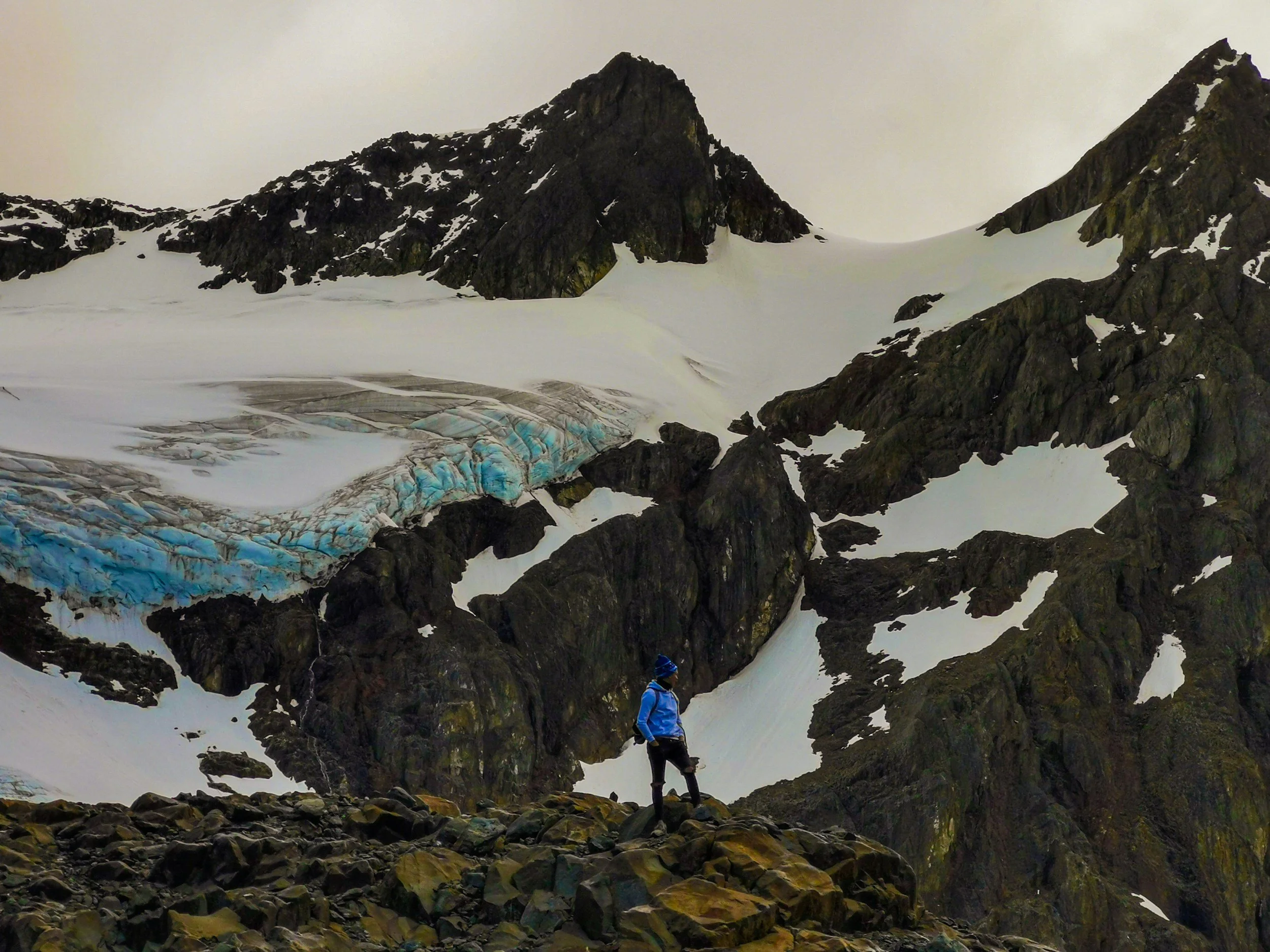 A hiker in a blue jacket and black pants standing on rocky terrain in front of snow-covered mountains and a glacier with blue ice.