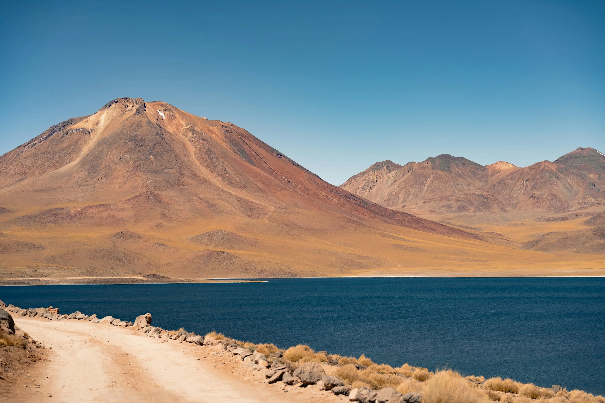 Scenic view of a mountain landscape with a large, brownish mountain and smaller mountains in the background, a blue lake in the foreground, and a dirt road along the lake's edge under a clear blue sky.