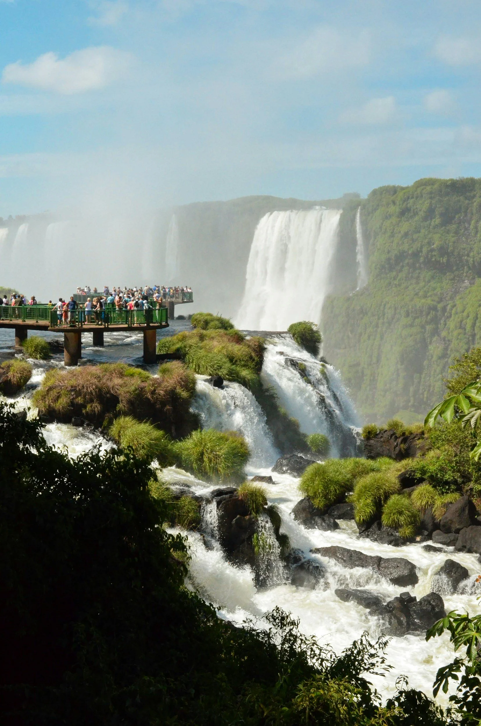 View of Niagara Falls with tourists on observation decks and green railings, surrounded by lush greenery and flowing water.