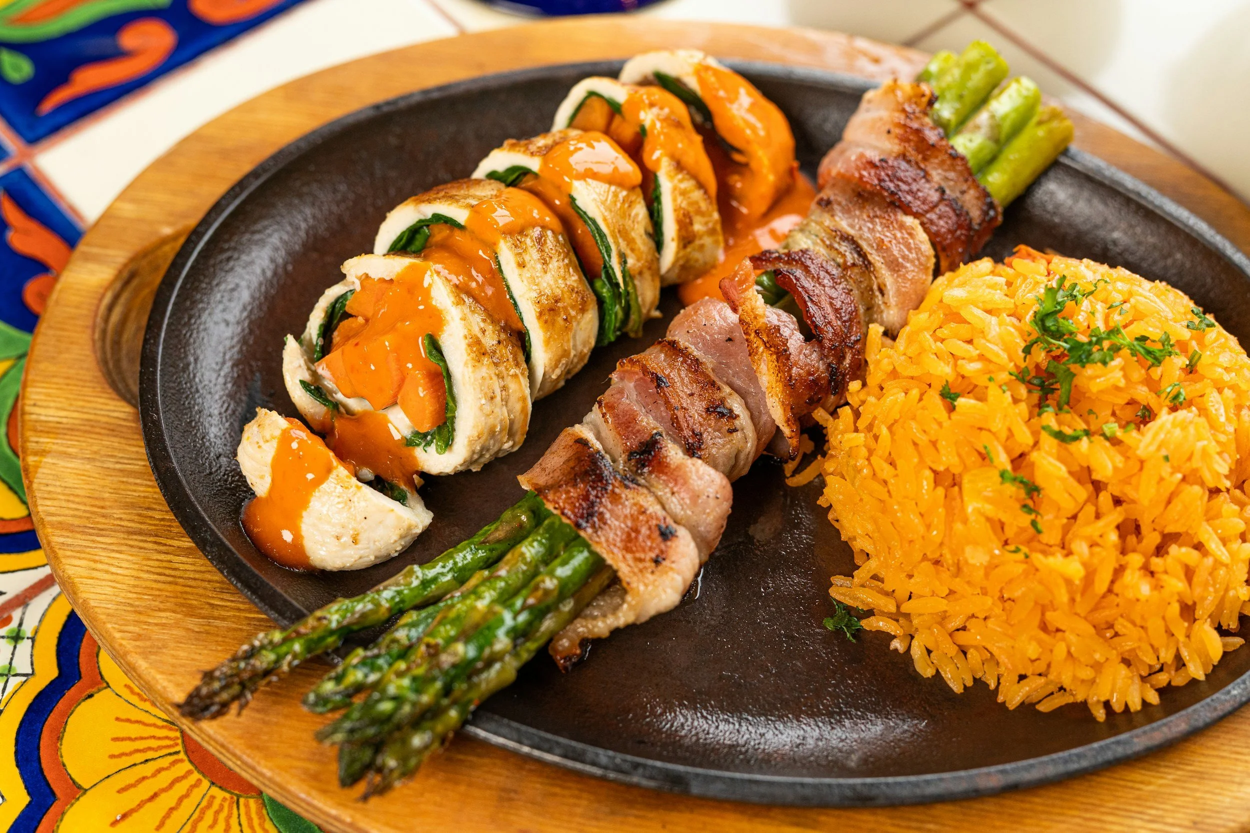 A sizzling skillet containing a chopped chicken and vegetable wrap, bacon-wrapped asparagus, and a mound of Mexican rice garnished with herbs.