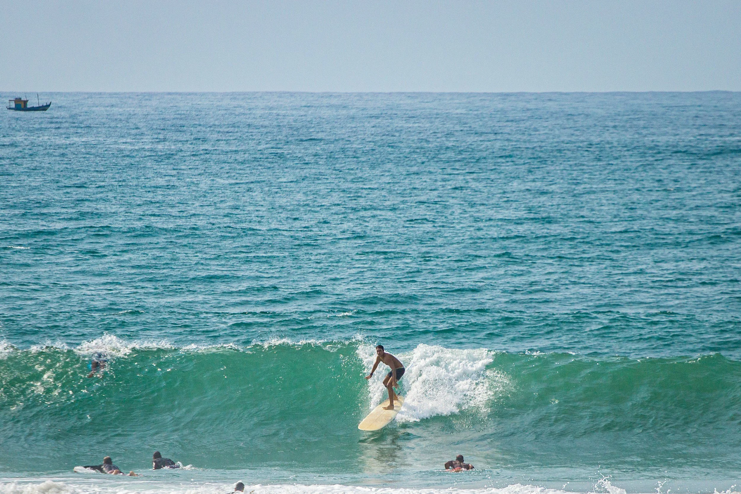 Joven surfer montando una ola en la playa con varias personas nadando y un barco en el mar al fondo.