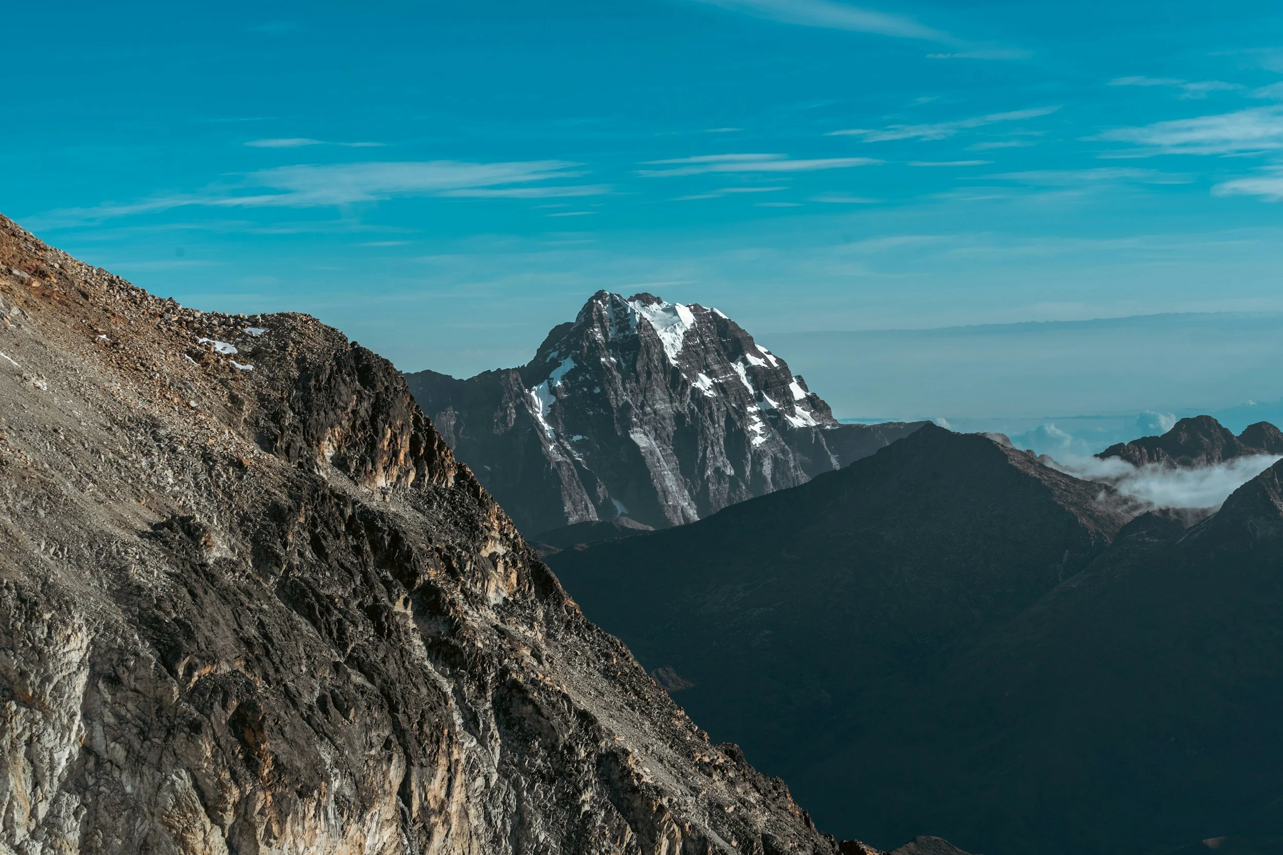 Majestic mountain landscape with a rugged, rocky foreground and a snow-capped peak in the distance under a blue sky with wispy clouds.