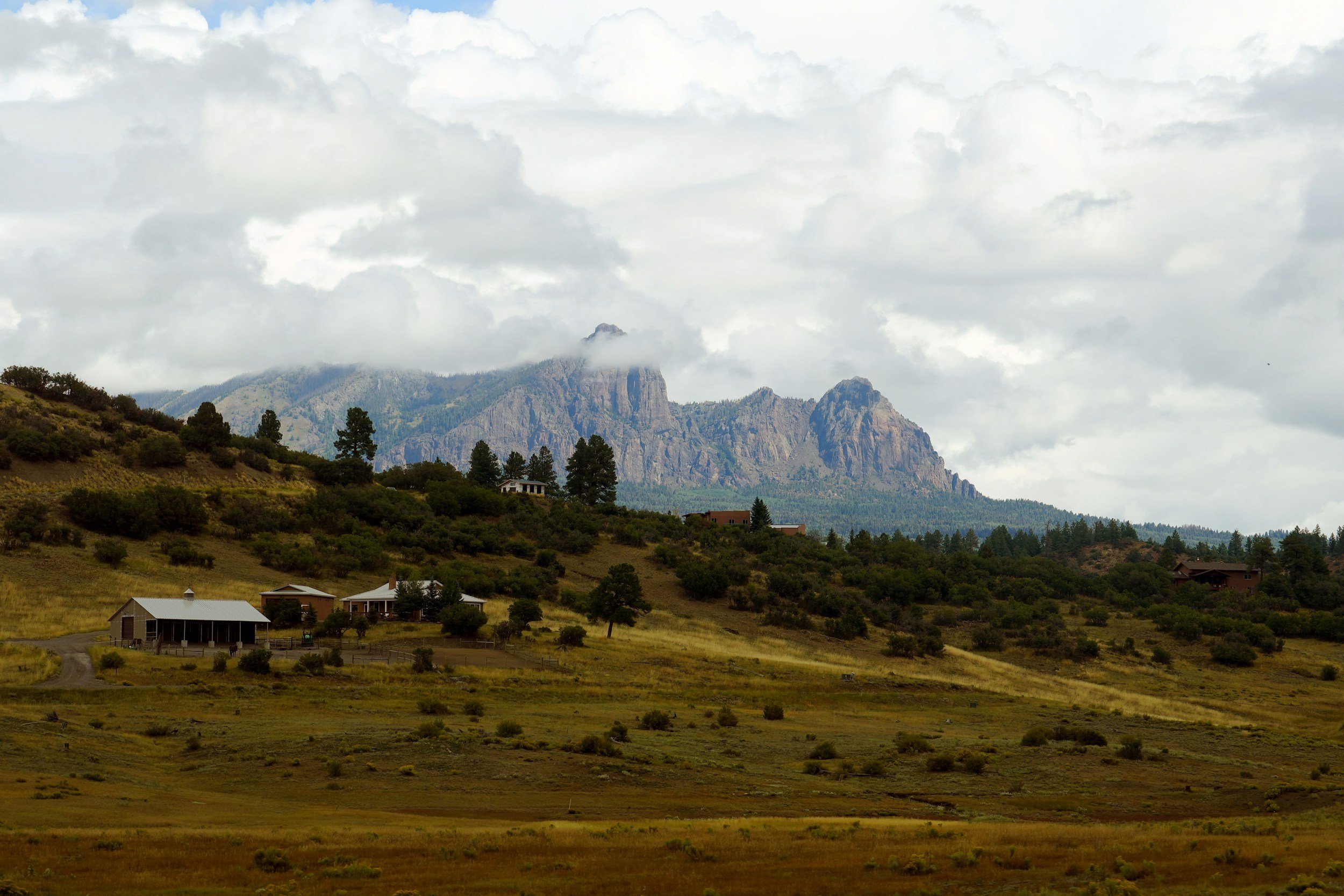 A scenic landscape of rolling hills with scattered houses, trees, and a mountain range in the background under partly cloudy skies.