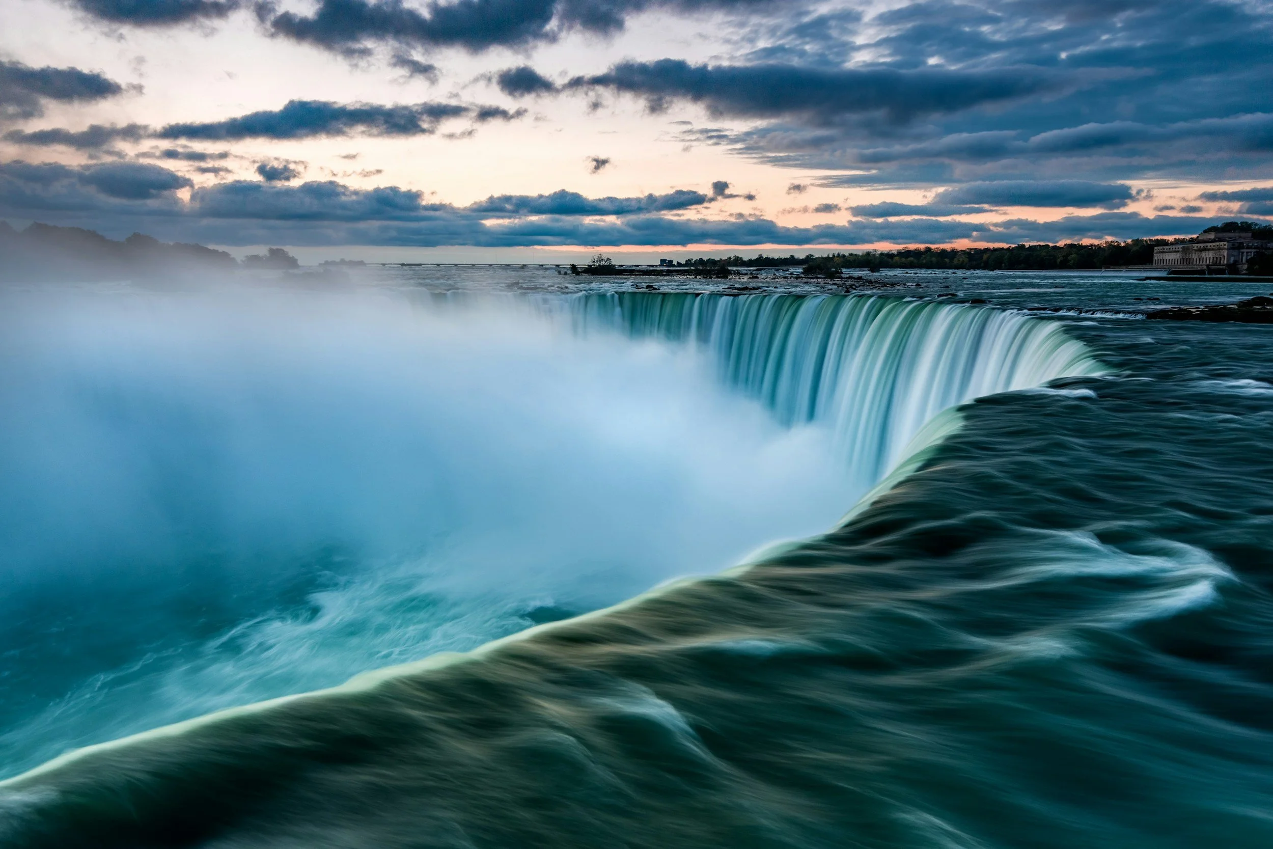 Niagara Falls at sunset with a cloudy sky, showing the waterfall with mist rising from the cascading water, surrounded by trees and buildings in the distance.
