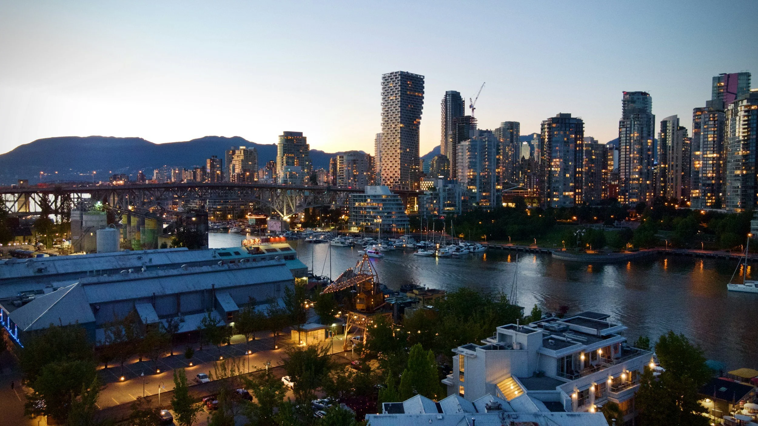 A city skyline at dusk with illuminated high-rise buildings, a bridge over a waterway, boats docked in the marina, and mountains in the background.