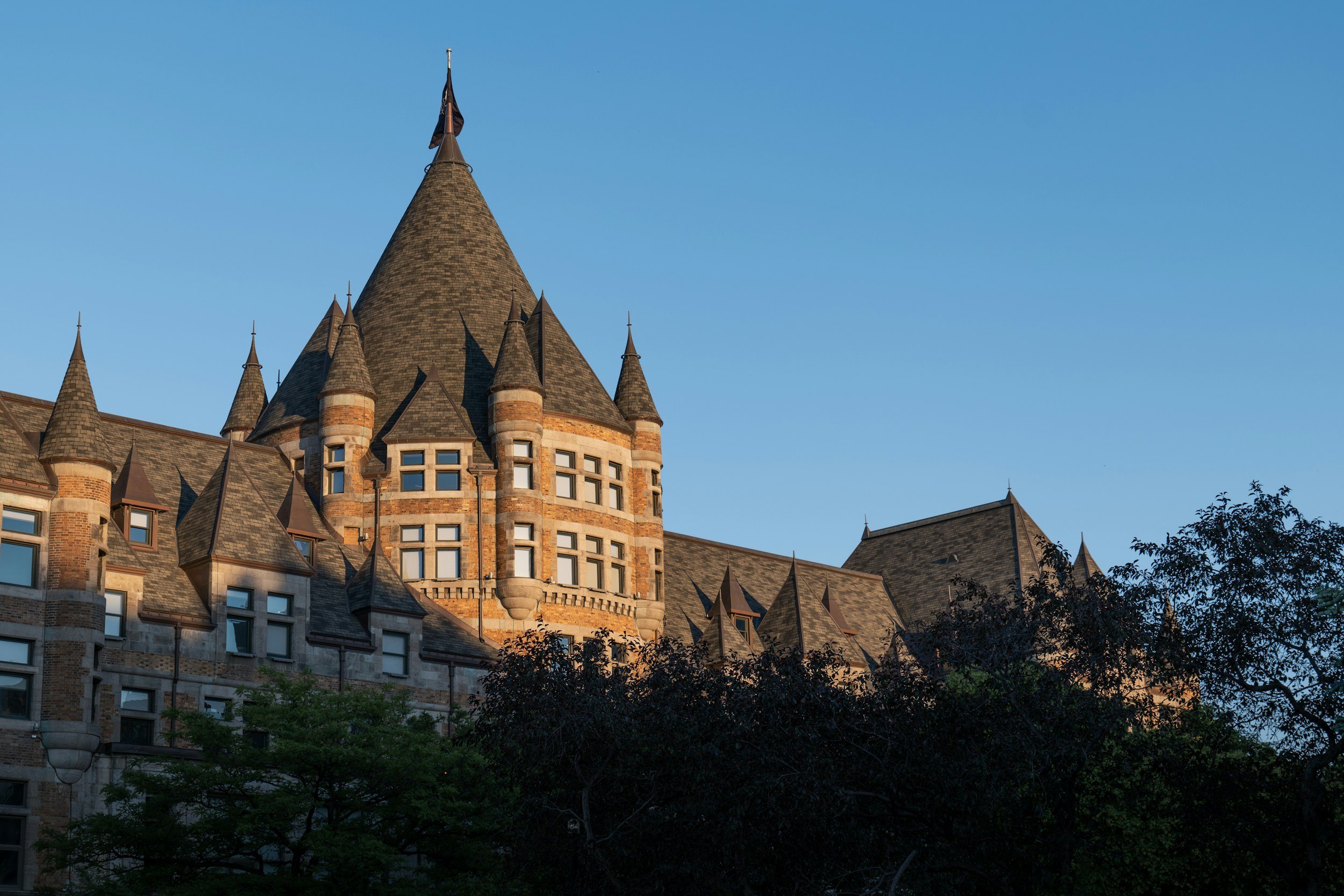 A historic castle-like building with multiple peaked roofs and a tall central tower, partially obscured by trees, under a clear blue sky.