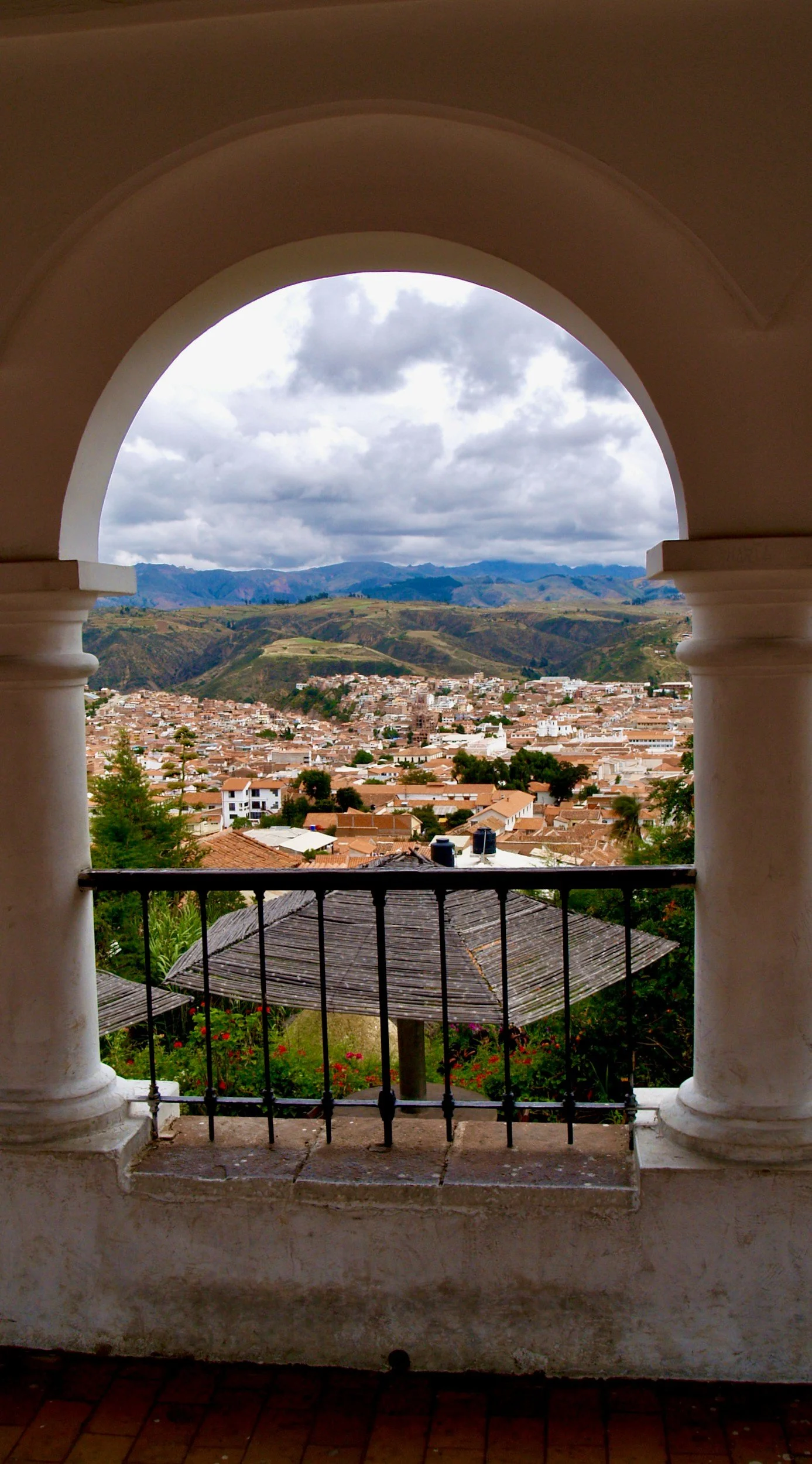 Scenic view of a town with terracotta roofs, mountains in the background, and cloudy sky, seen through an arched window with white columns and black railing.