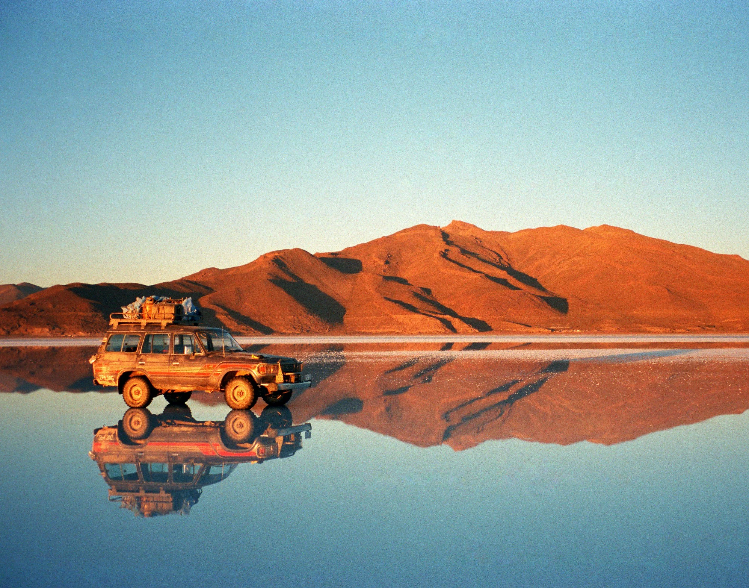 A SUV driving on flat terrain with a mountain range in the background during sunset, and a reflection of both the vehicle and mountains in a body of water.